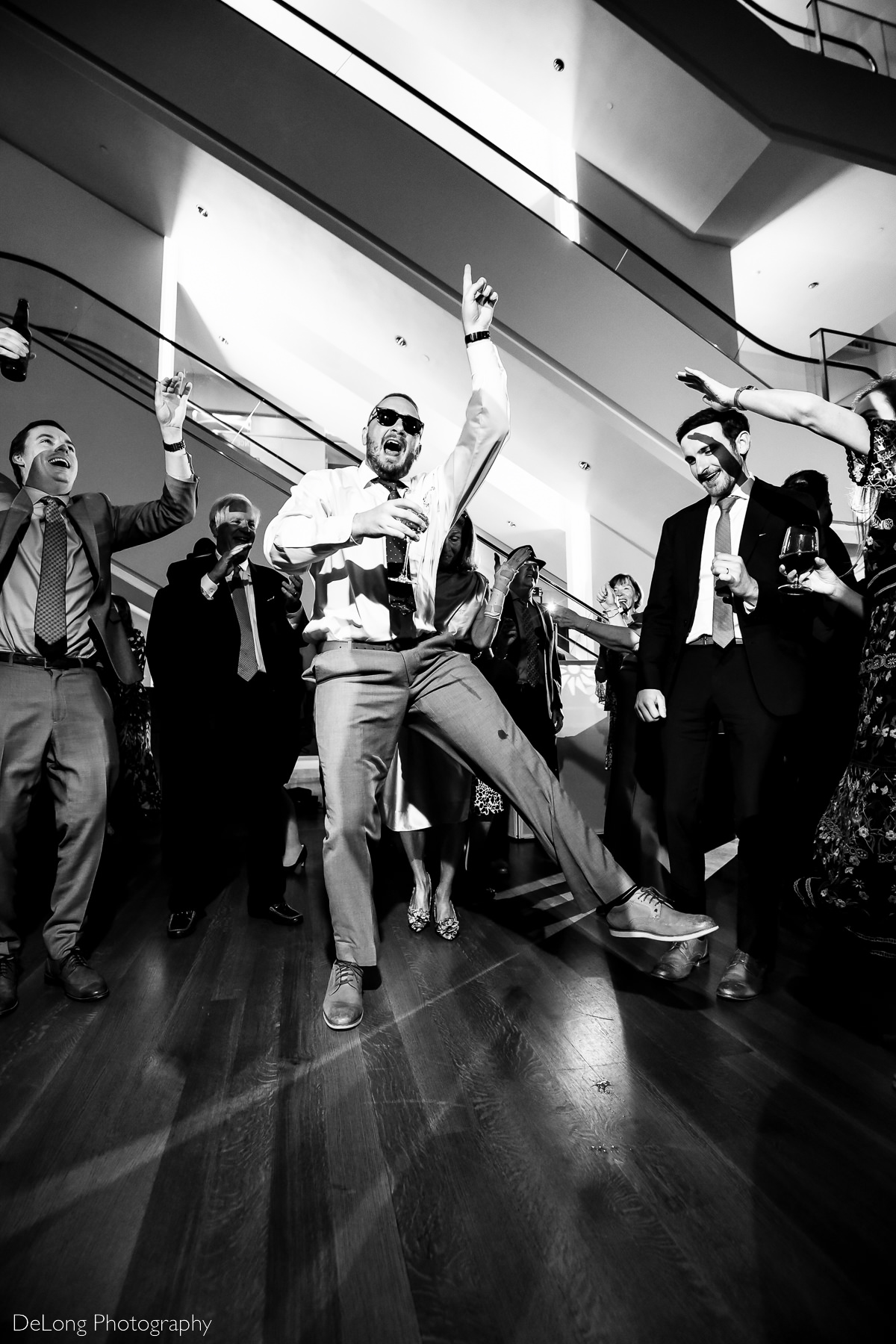 Black and white image of guest in sunglasses holding a drink and jumping on the dance floor of the Mint Museum Uptown by Charlotte wedding photographers DeLong Photography