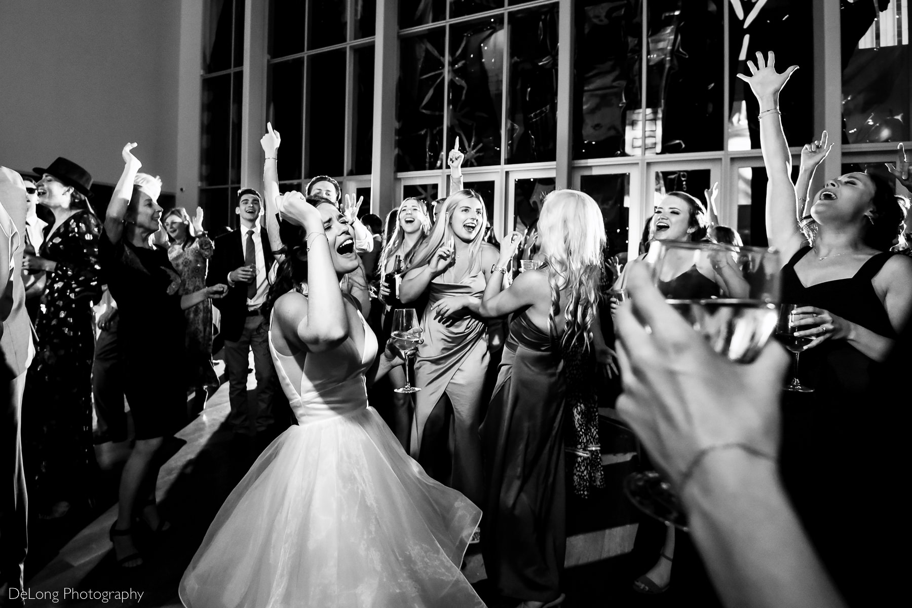 Black and white image of bride singing loudly with her eyes closed surrounded by other singing guests with their hands in the air on the dance floor of the Mint Museum by Charlotte wedding photographers DeLong Photography