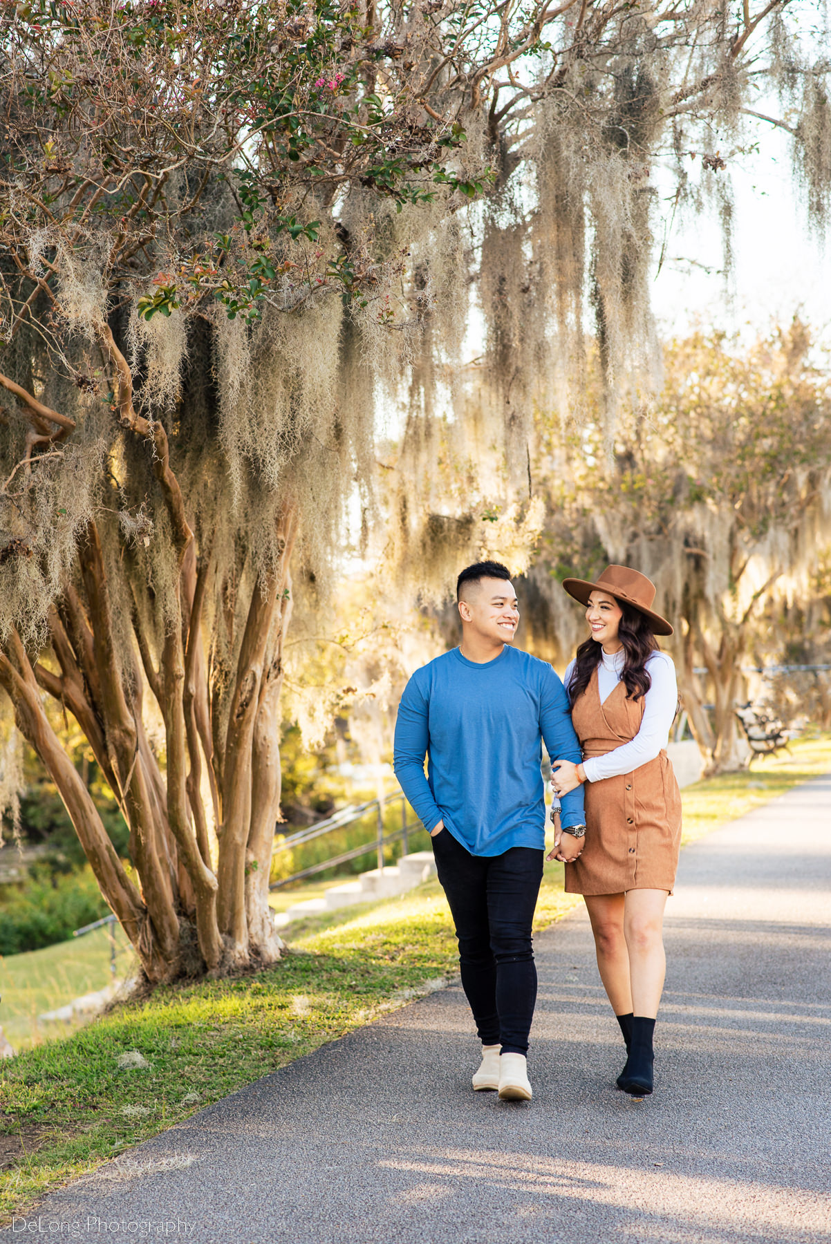 newly engaged couple walking among he mossy trees on sidewalk and smiling at one another at Riverfront Park in Columbia, SC. Photo by Charlotte wedding photographers DeLong Photography.