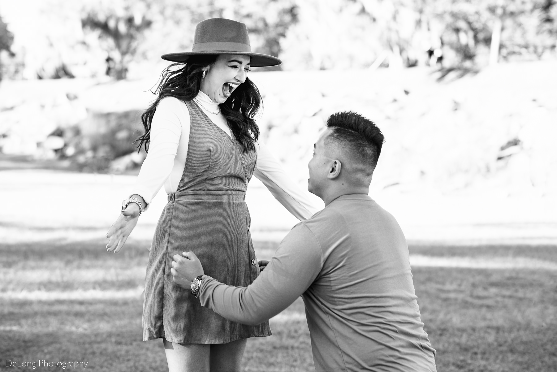 Black and white image of woman holding out arms for a hug and joyful reaction to surprise proposal at Riverfront Park in Columbia, SC. Photo by Charlotte wedding photographers DeLong Photography.