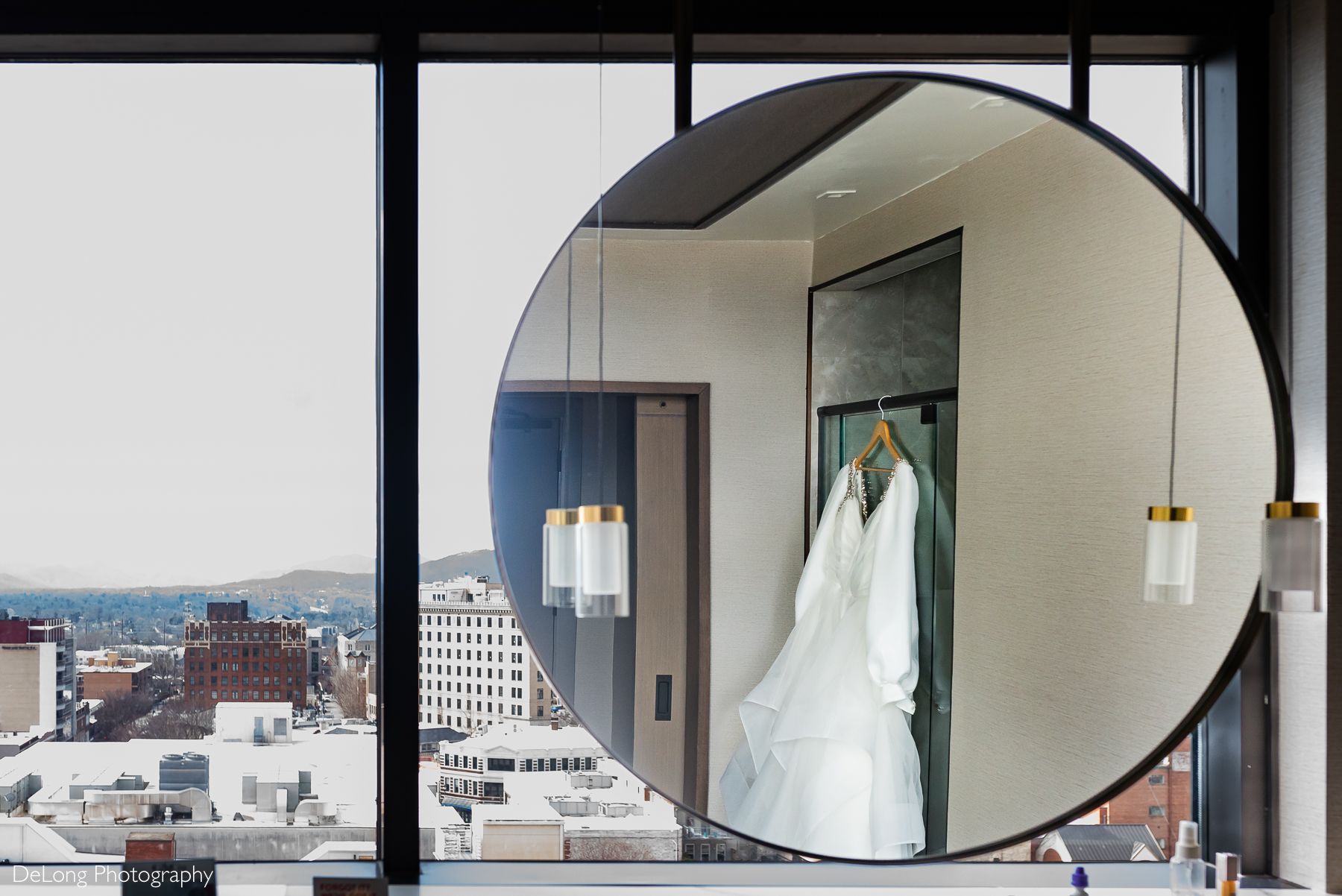 Wedding dress reflected in a circular mirror, with the city of Asheville visible through large glass windows in the background. Photograph by Charlotte wedding photographers DeLong Photography taken in the Kimpton Hotel Arras.