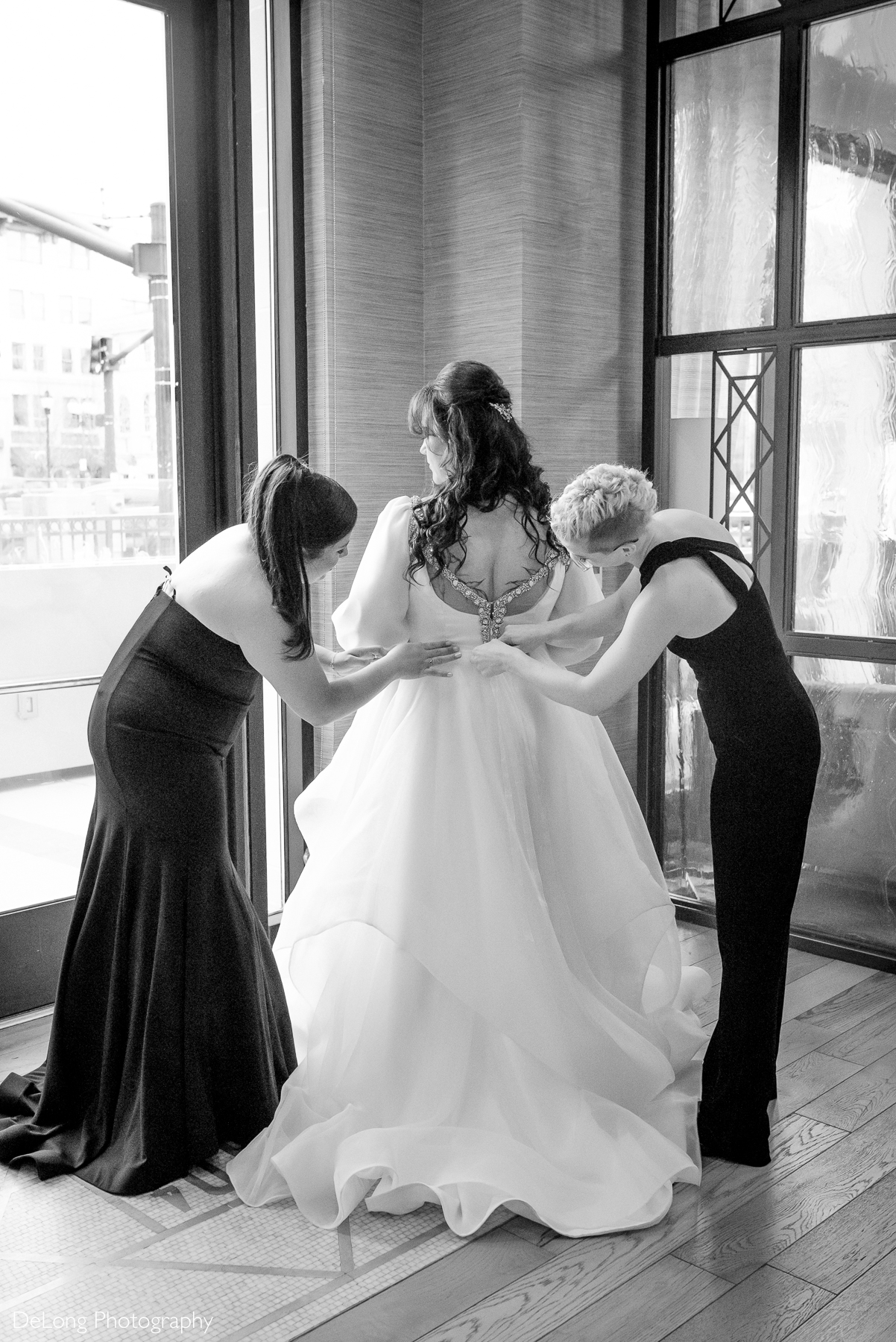 Black and white candid photograph of the bride having her dress zipped and adjusted by her maids of honor. Photograph by Charlotte wedding photographers DeLong Photography taken in the Kimpton Hotel Arras.