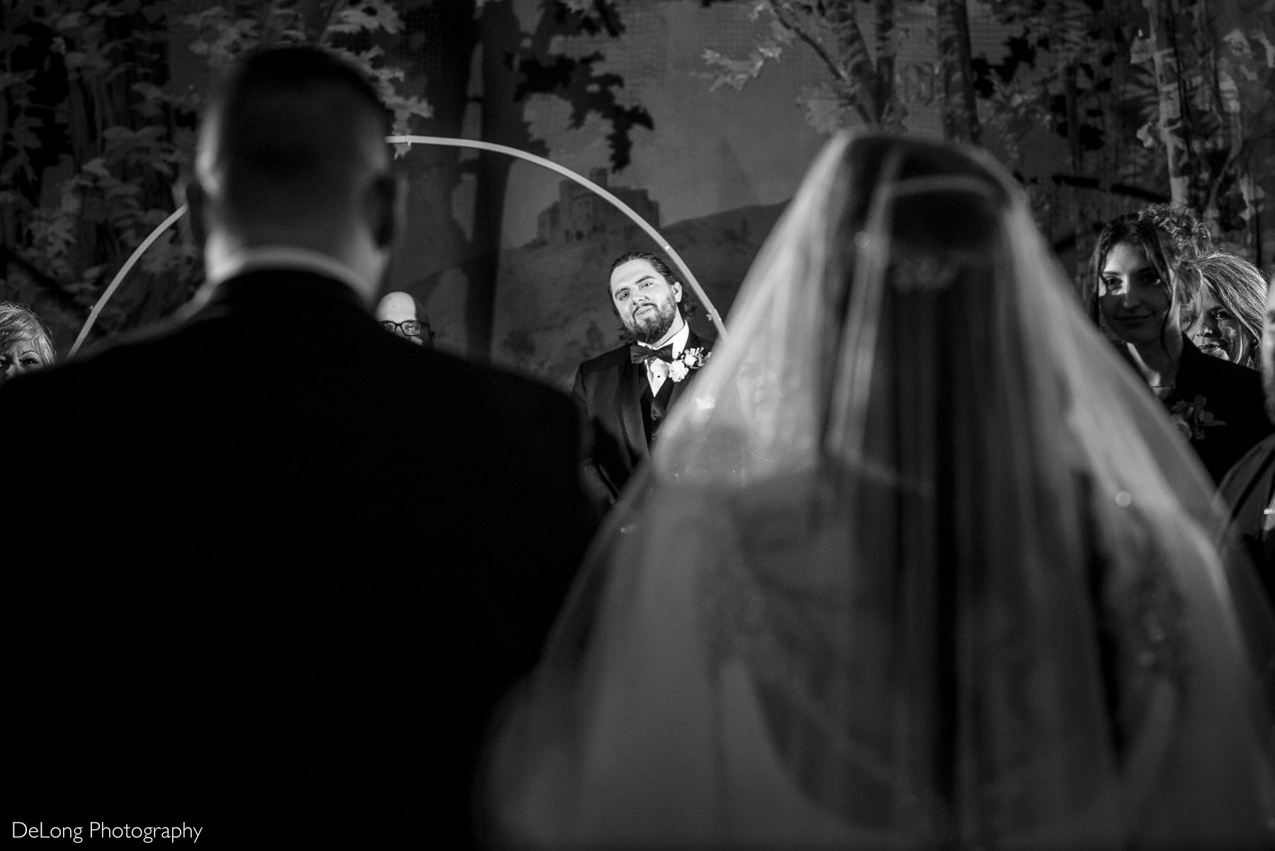 Groom smiling look at his bride coming down the aisle.The groom is framed between the bride and her escort as the make their way down the aisle. Photograph by Charlotte wedding photographers DeLong Photography taken at the Asheville Masonic Temple.