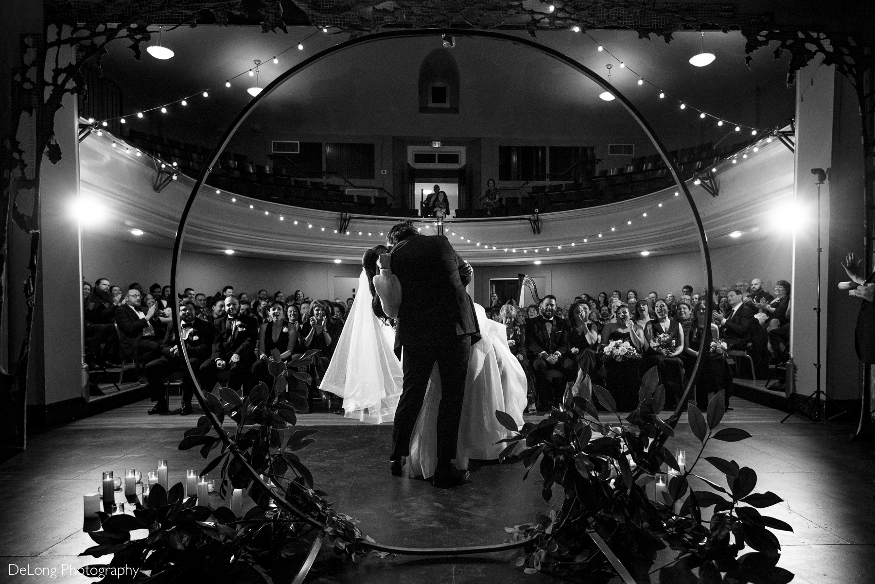 Black and white photograph of the groom kissing his bride, dipping her. The viewpoint is on stage facing the guests. Photograph by Charlotte wedding photographers DeLong Photography taken at the Asheville Masonic Temple.