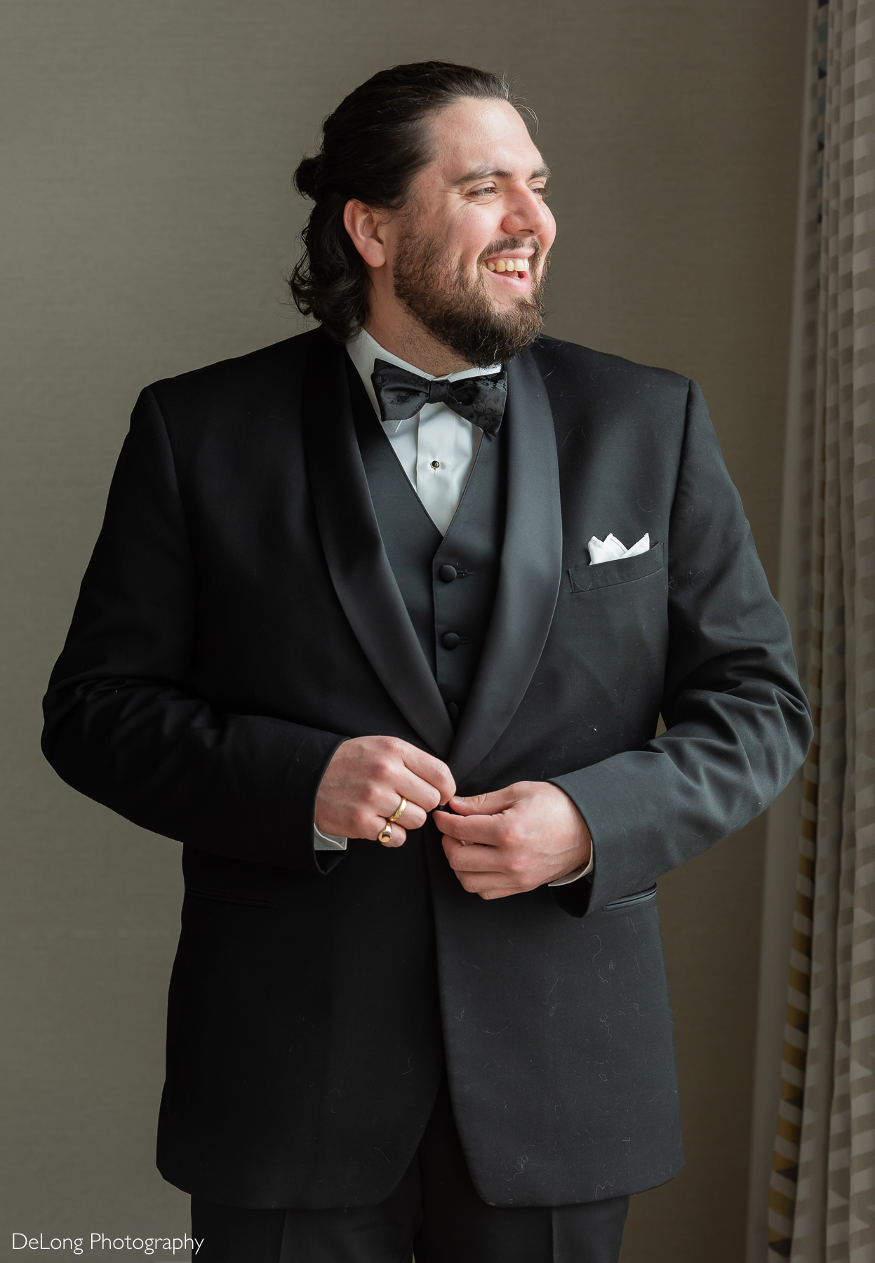 Groom laughing as he buttons his jacket and looks out the window. Photograph by Charlotte wedding photographers DeLong Photography taken in the Kimpton Hotel Arras.