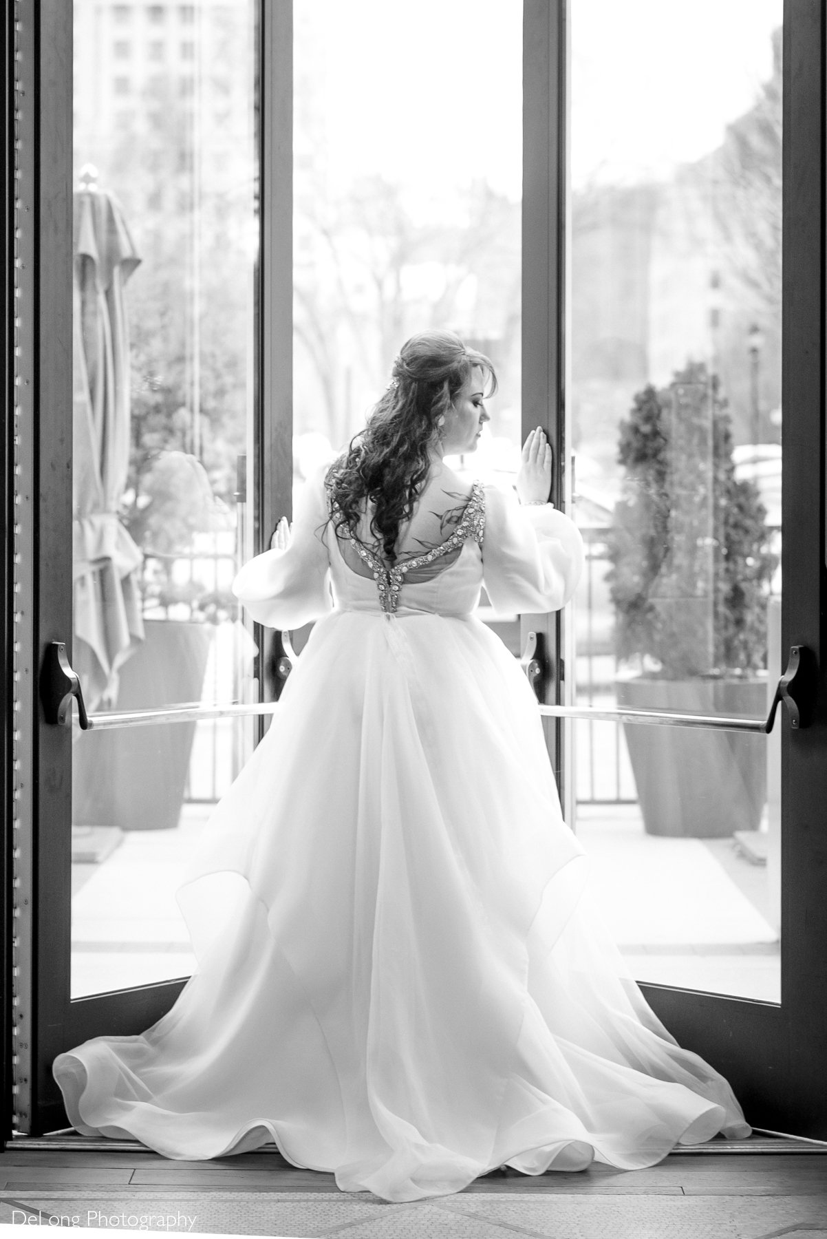 Black and white bridal portrait of the bride opening the doors of the downstairs bar of the Kimpton Hotel Arras. The image showcases the jeweled straps of her wedding dress along with her back tattoos that mimic the dress design. Photograph by Charlotte wedding photographers DeLong Photography.