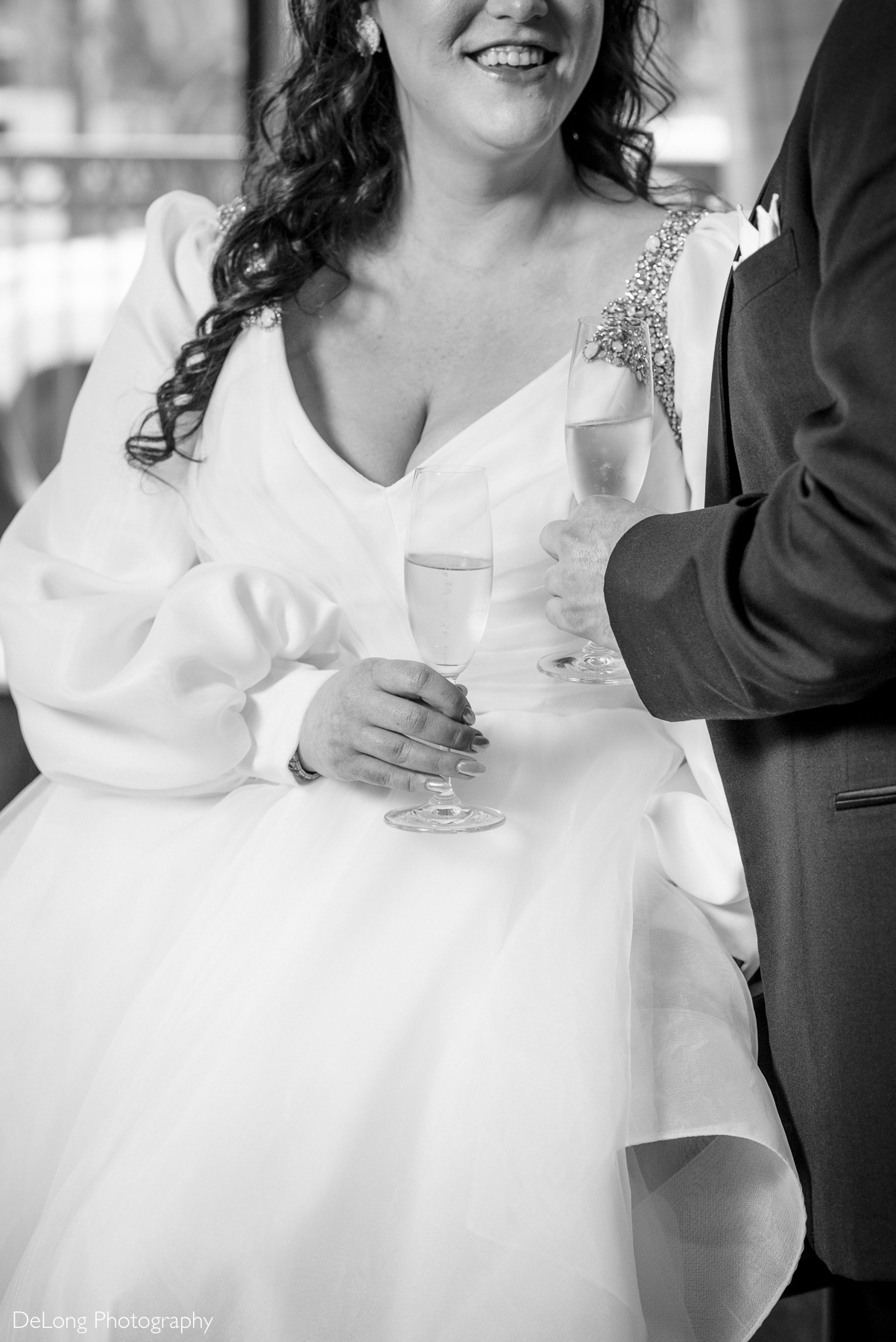 Black and white photograph cropped so it only includes the bride's smile and the champagne glasses the couple is holding. Groom smiling at his bride after turning around and seeing her for the first time during their first look. Photograph by Charlotte wedding photographers DeLong Photography taken in the Kimpton Hotel Arras.