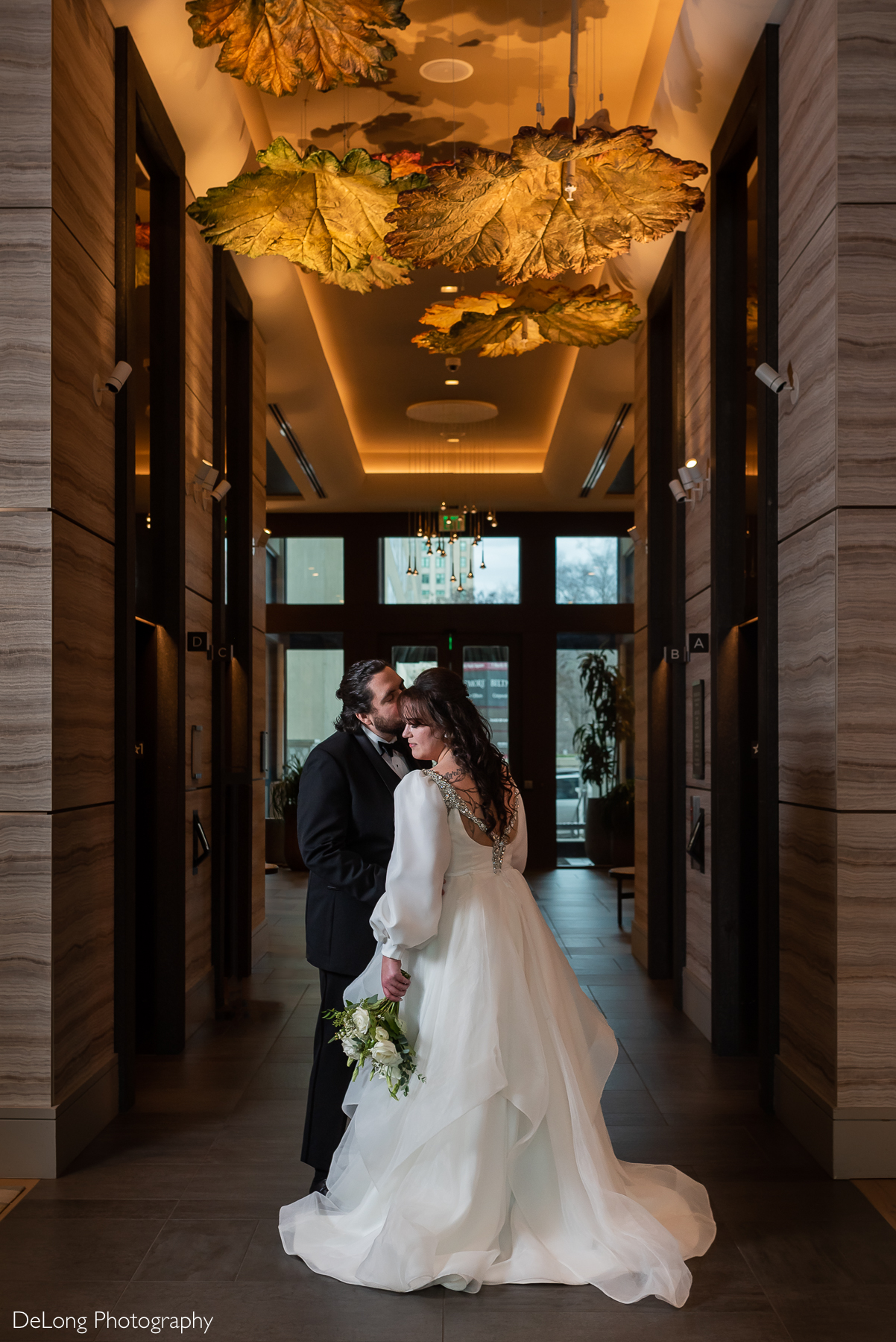 Formal portrait of the groom kissing his bride's temple underneath the leaf installation in the hotel lobby. Photograph by Charlotte wedding photographers DeLong Photography taken in the Kimpton Hotel Arras.