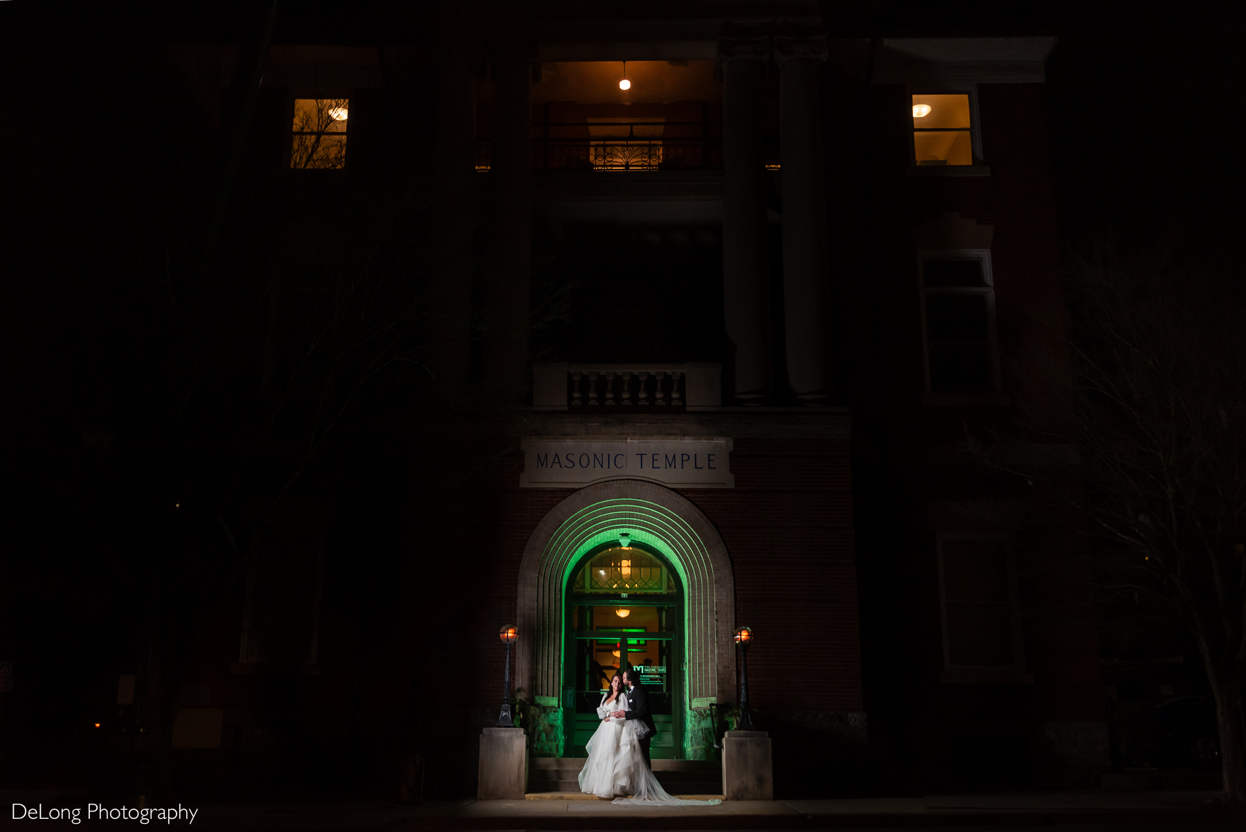 Night portrait of the couple, small in the frame, backlit with a green gel, outside the Asheville Masonic Temple. Photograph by Charlotte wedding photographers DeLong Photography.