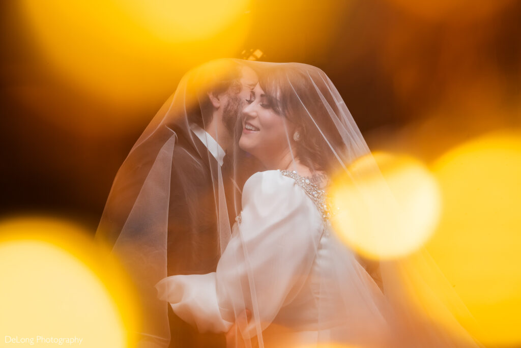 A bride smiles brightly as her groom kisses her on the cheek under her flowing veil. The couple is outside the Asheville Masonic Temple at night, surrounded by a dreamy ambiance with soft, glowing bokeh lights in the foreground. This romantic moment was beautifully captured by DeLong Photography