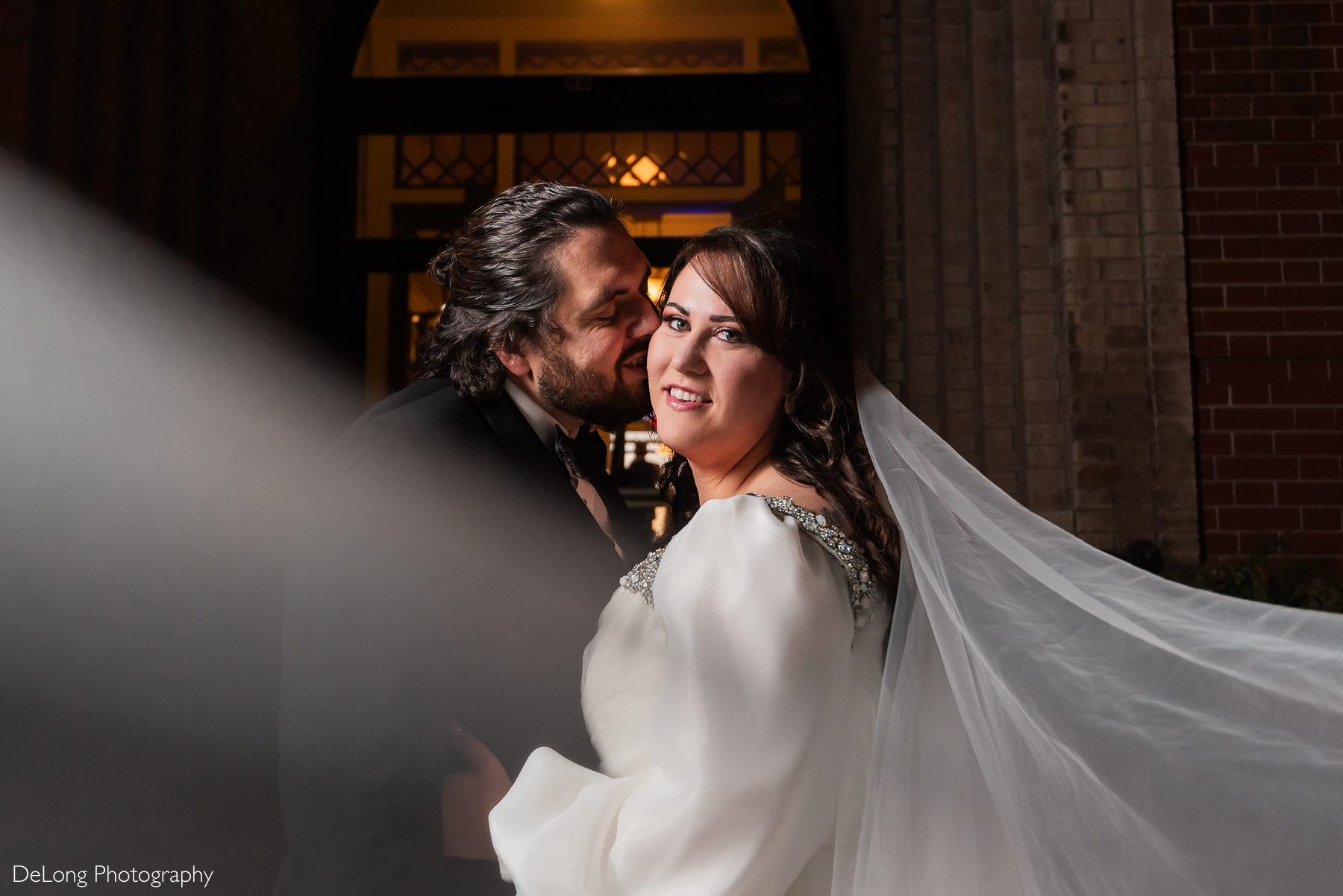 Bride looking at the camera while the groom kisses her cheek. Her veil swoops from her hair to the left side of the frame. Photograph by Charlotte wedding photographers DeLong Photography taken at the Asheville Masonic Temple.