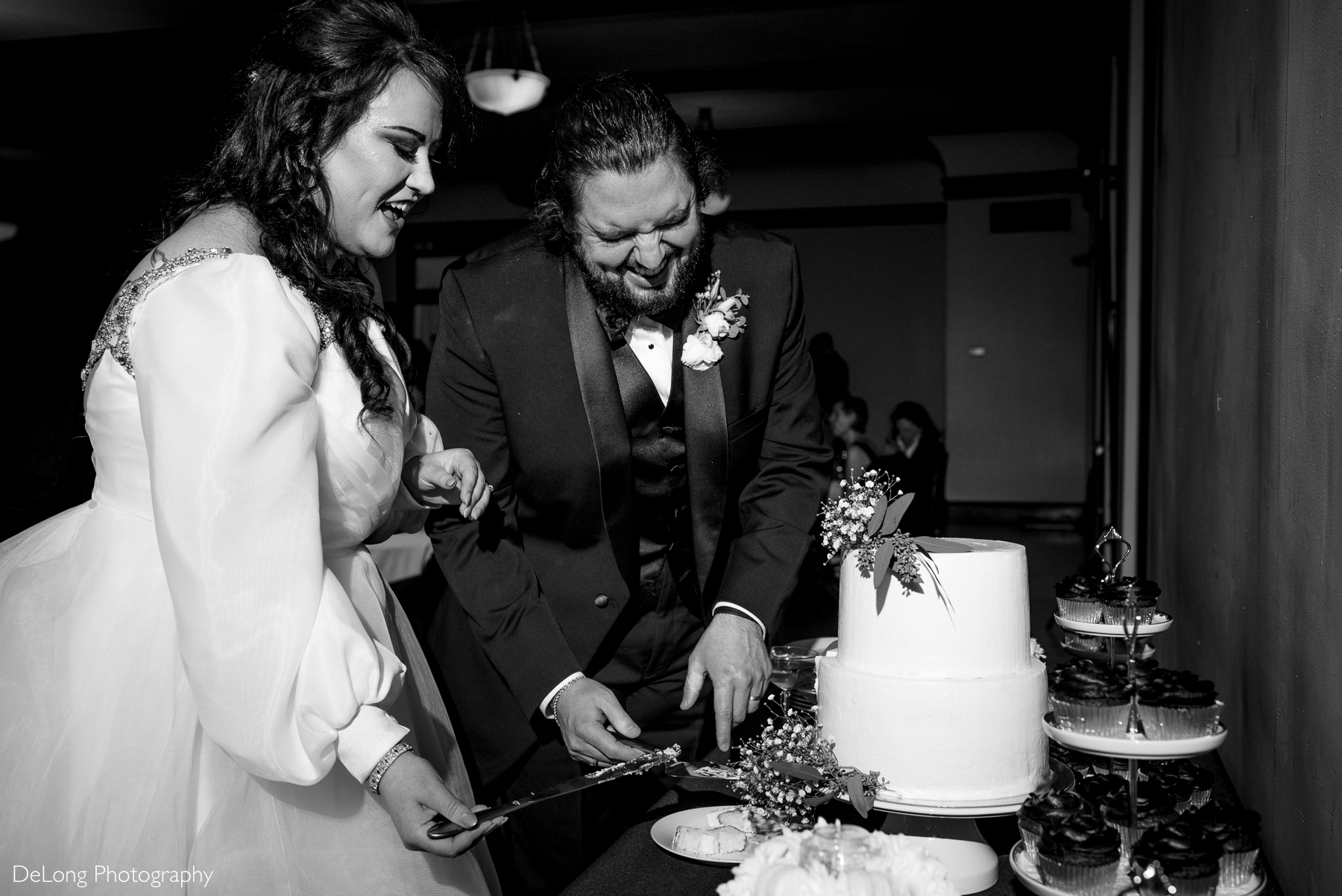 Black and white image of groom cracking up laughing while he and his bride cut their wedding cake. Photograph by Charlotte wedding photographers DeLong Photography taken at the Asheville Masonic Temple.