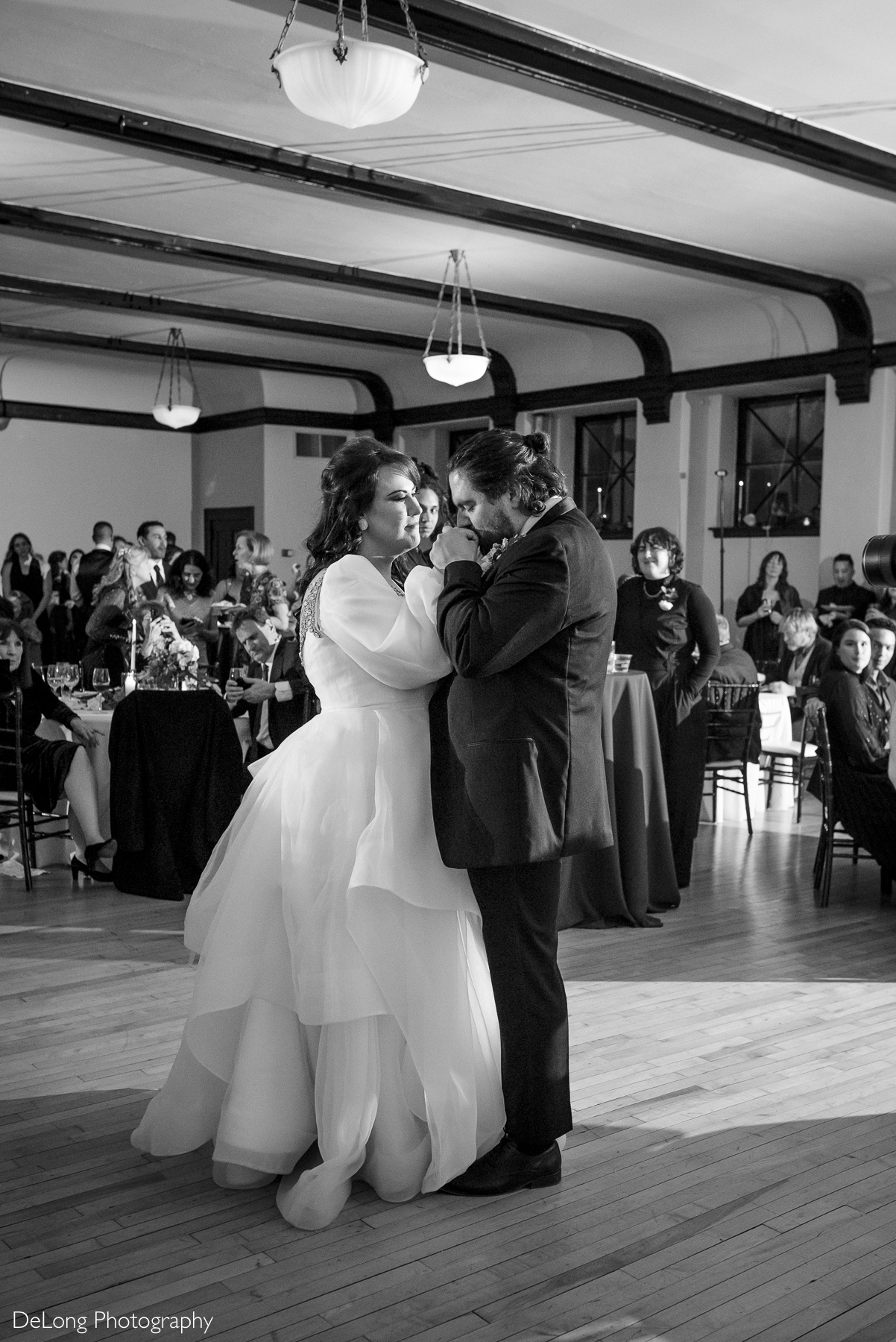 Black and white photograph of an emotional bride as the groom kisses her hang during their first dance. Photograph by Charlotte wedding photographers DeLong Photography taken at the Asheville Masonic Temple.