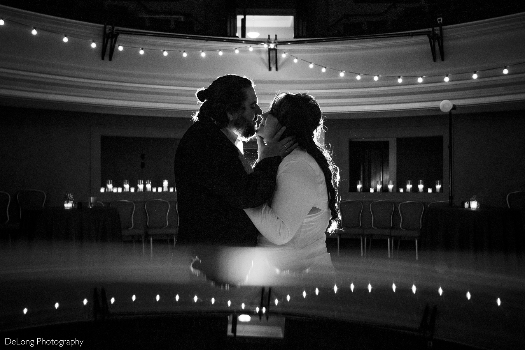 Black and white photograph in the auditorium of the Asheville Masonic Temple of the groom bringing his bride in for a kiss, his hands cupping her face. The fairy lights handing from the ceiling are mirrored in the bottom of the frame. Photograph by Charlotte wedding photographers DeLong Photography.