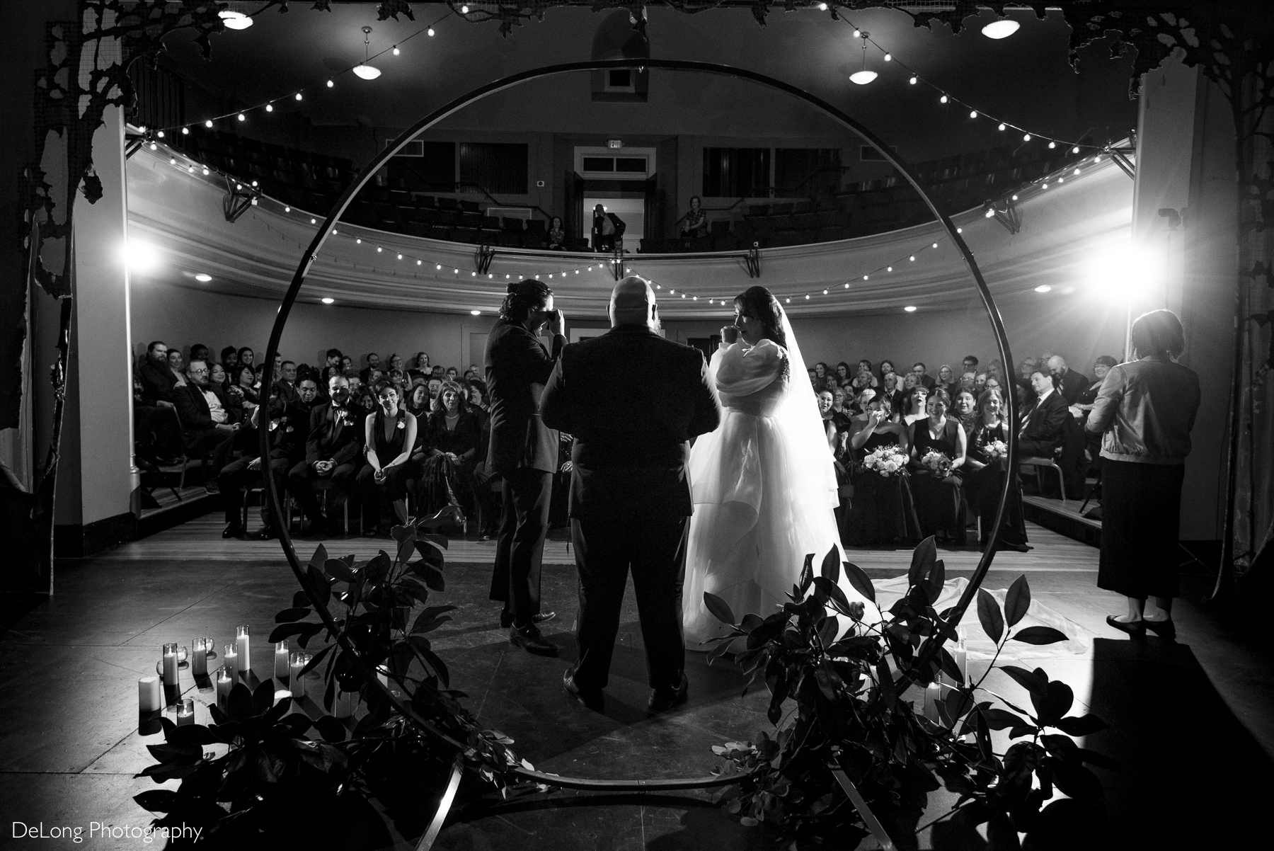 Candid black and white image of the bride and groom wiping tears during their wedding ceremony. The viewpoint is from on stage facing the guests. Photograph by Charlotte wedding photographers DeLong Photography taken at the Asheville Masonic Temple.