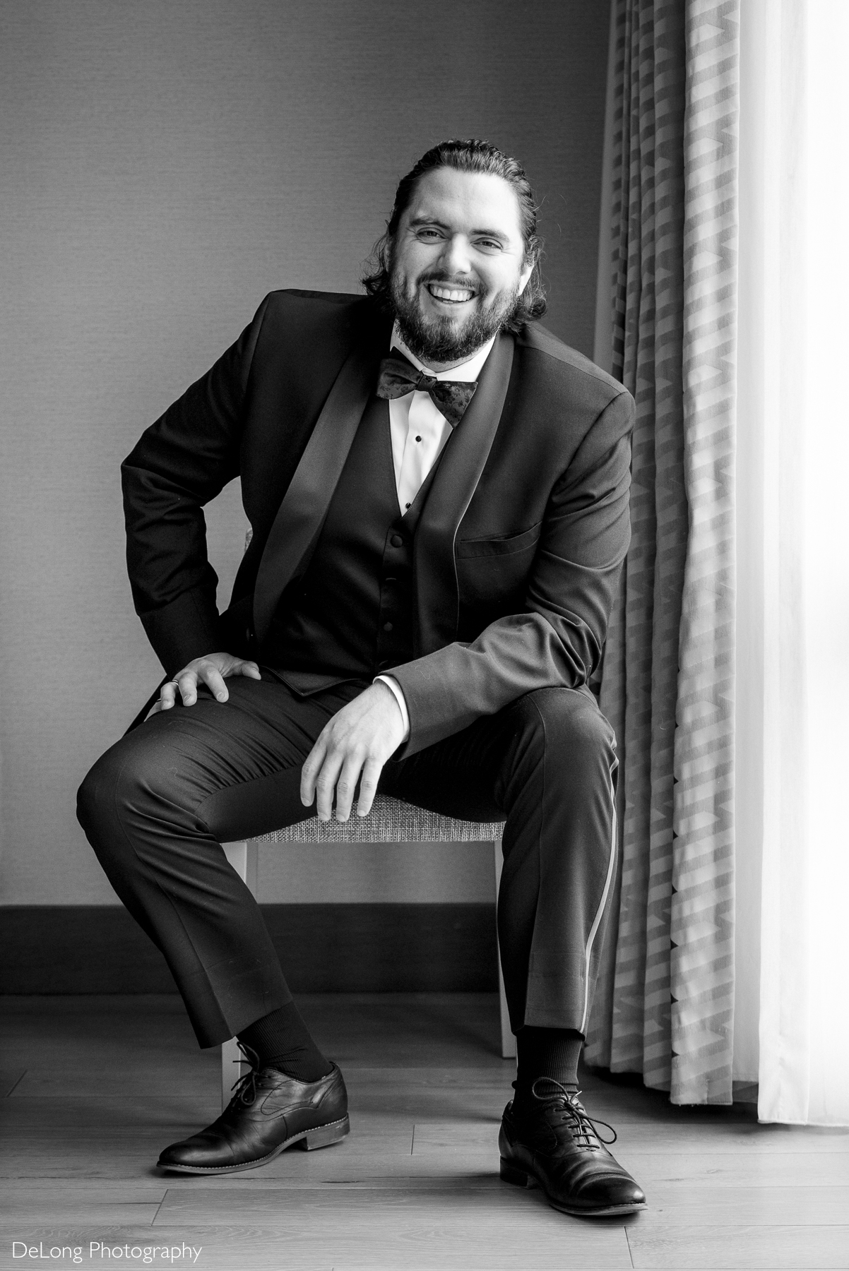 Black and white portrait of the groom seated, leaning on one forearm, smiling while looking at the camera. Photograph by Charlotte wedding photographers DeLong Photography taken in the Kimpton Hotel Arras.