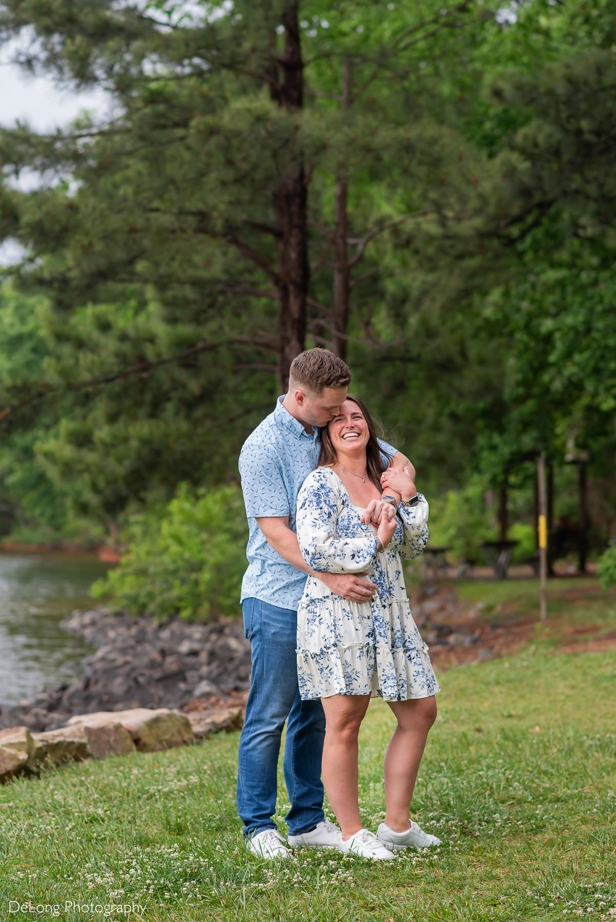 Man kisses his fiancée's temple, his arms around her. She is smiling, hugging his arms close. Photo taken in the grass near the water's edge at Jetton Park.