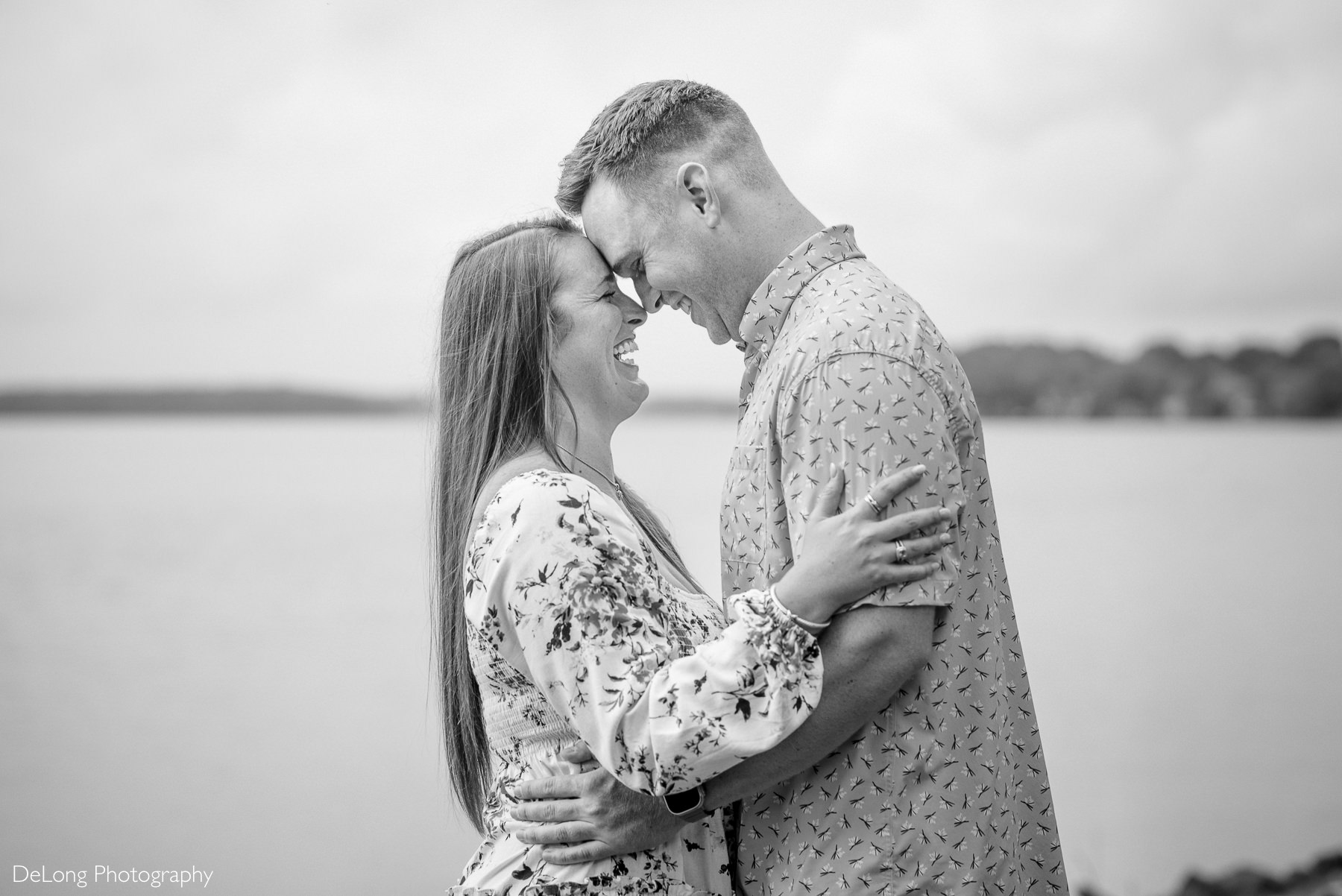 Black and white photo of a newly engaged couple smiling with their foreheads together with the water of Jetton Park in the background.