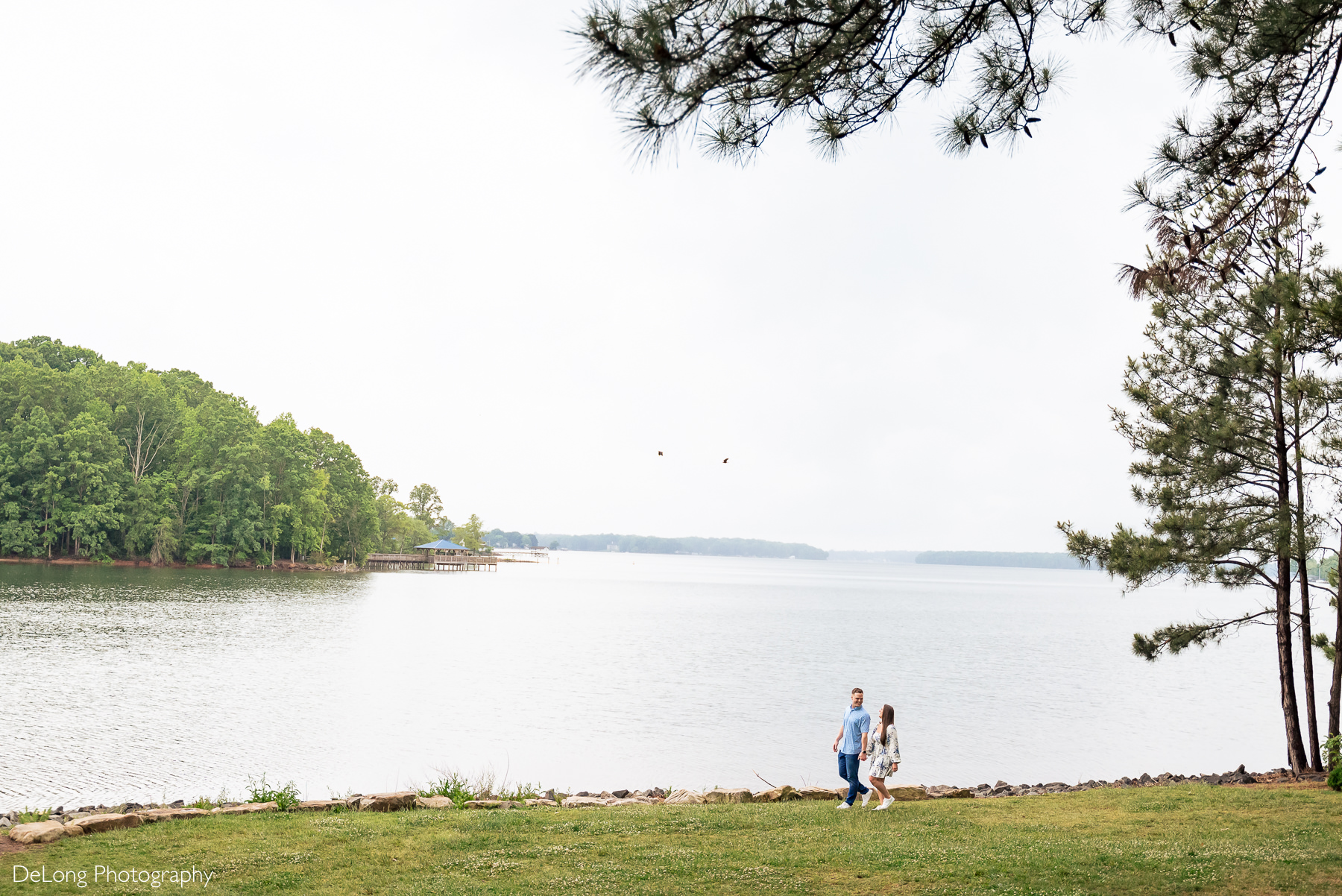 Wide angle view of the lake at Jetton Park with a newly engaged couple, small in the frame, walk from right to left along the shoreline smiling at one another.