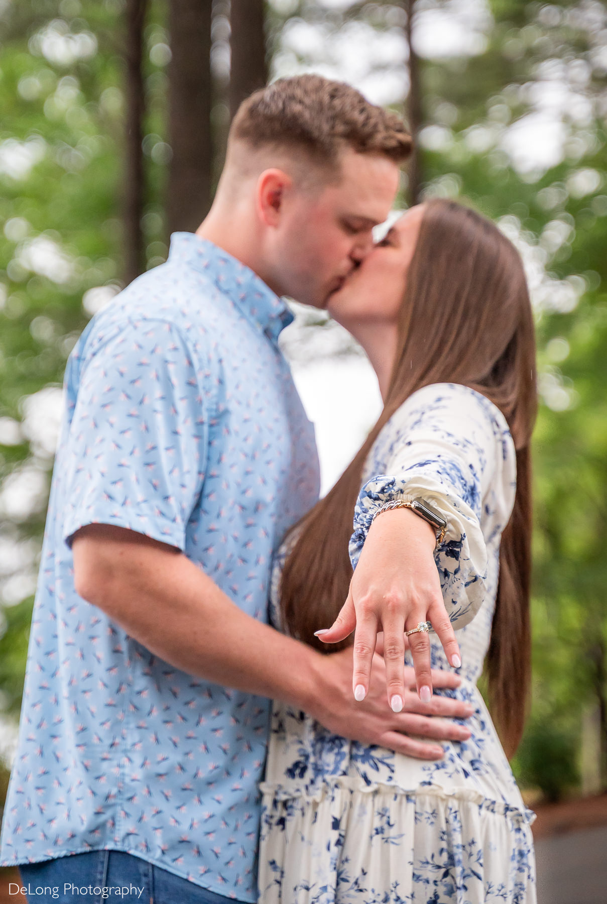 Focus is on the engagement ring on the woman's hand. She is holding it out toward the camera while the couple us blurry, sharing a kiss, in the background.