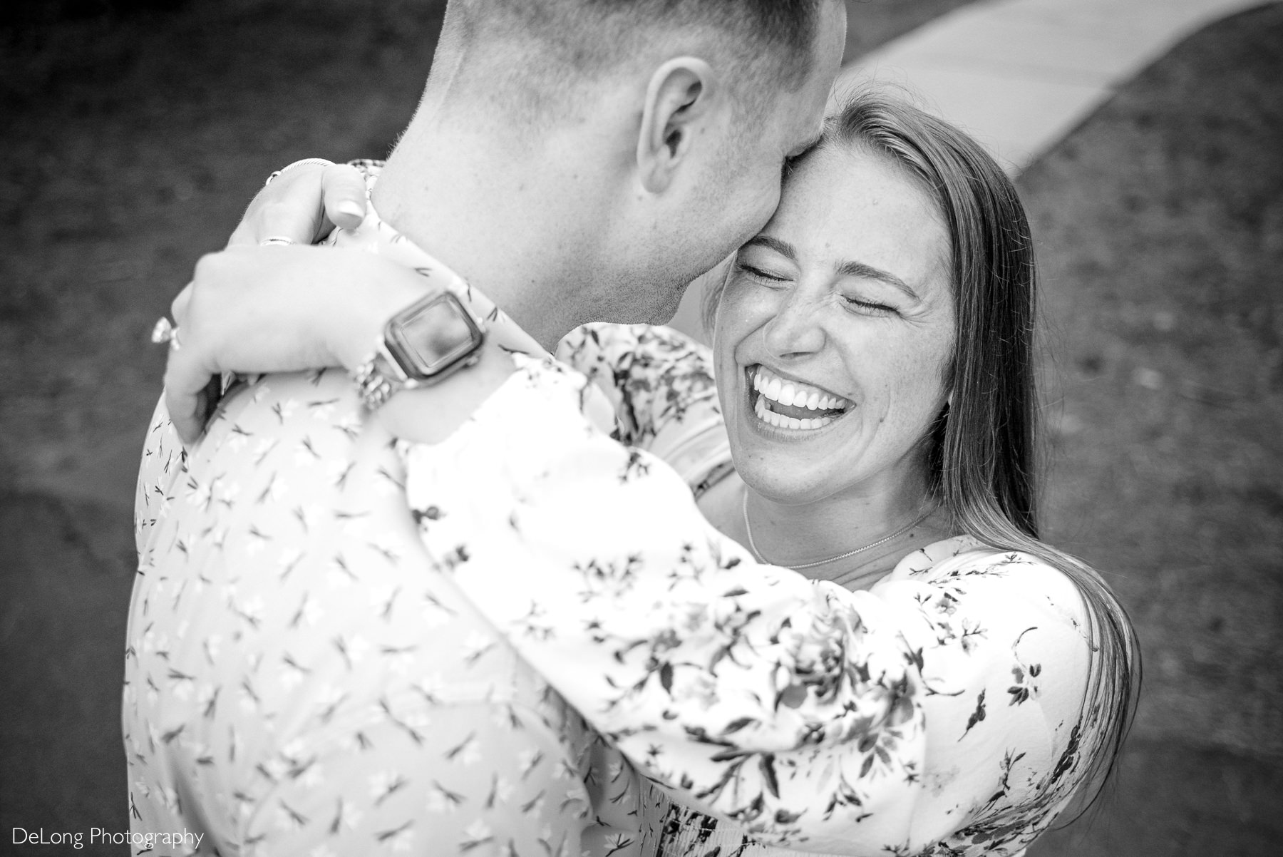 Top down candid photograph taken over the man's shoulder. His fiancée has her arms around his neck and is cracking up laughing as he whispers in her ear.