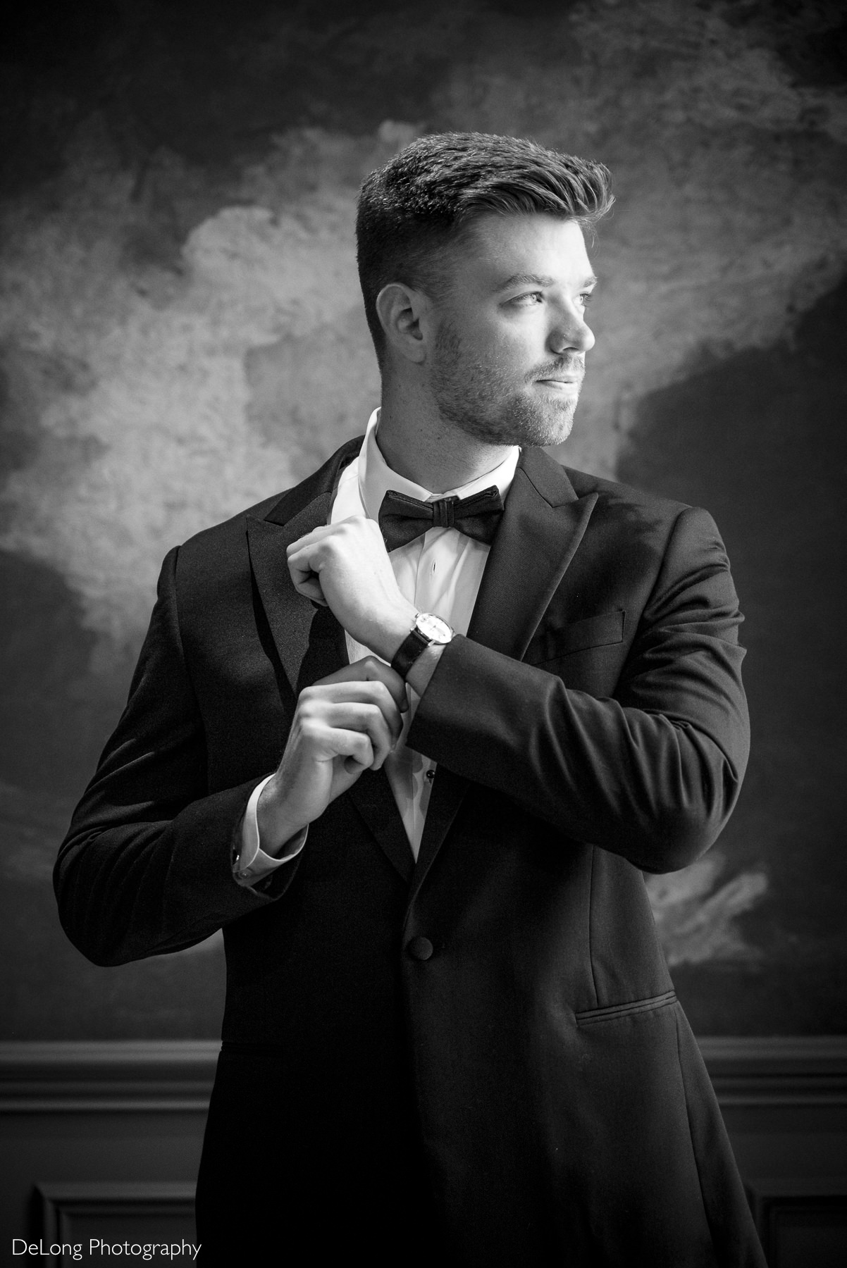 A black-and-white photograph of a groom getting ready at Carmel Country Club. The groom, dressed in a classic black tuxedo with a bow tie, has a calm and confident expression as he looks out the window and us adjusting his watch band. The moody lighting and textured background add a timeless and elegant feel to the moment. Photographed by DeLong Photography.
