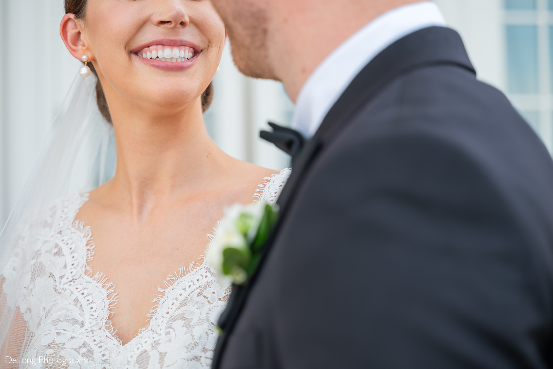 A close-up of a bride smiling joyfully at her groom on their wedding day at Carmel Country Club. The intricate lace details of her gown and pearl earrings complement her radiant expression, while the groom, partially visible in a tuxedo, adds depth to the intimate moment. Photograph by DeLong Photography.
