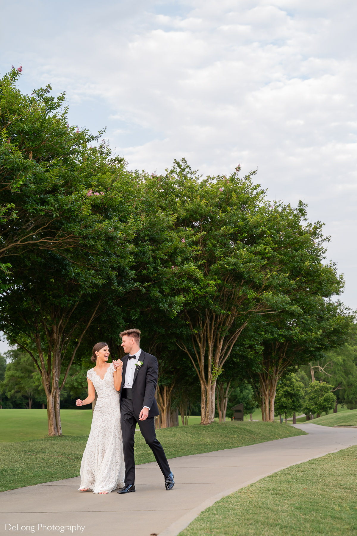 A newlywed couple walks on the tree-lined sidewalk along the golf course of Carmel Country Club. They are holding hands and giggling. The bride wears an elegant lace gown, while the groom looks dapper in a classic black tuxedo. Captured by DeLong Photography.