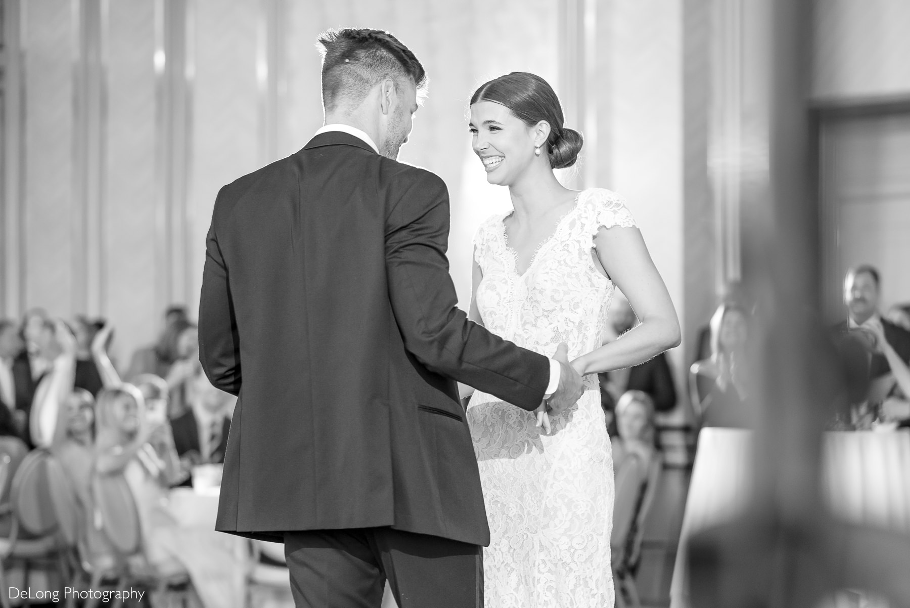 A black-and-white photograph of a bride laughing, handing hands with her groom at the end of their first dance at Carmel Country Club. The bride wears an elegant lace gown with cap sleeves, while the groom looks dapper in a classic black tuxedo.Captured by DeLong Photography.