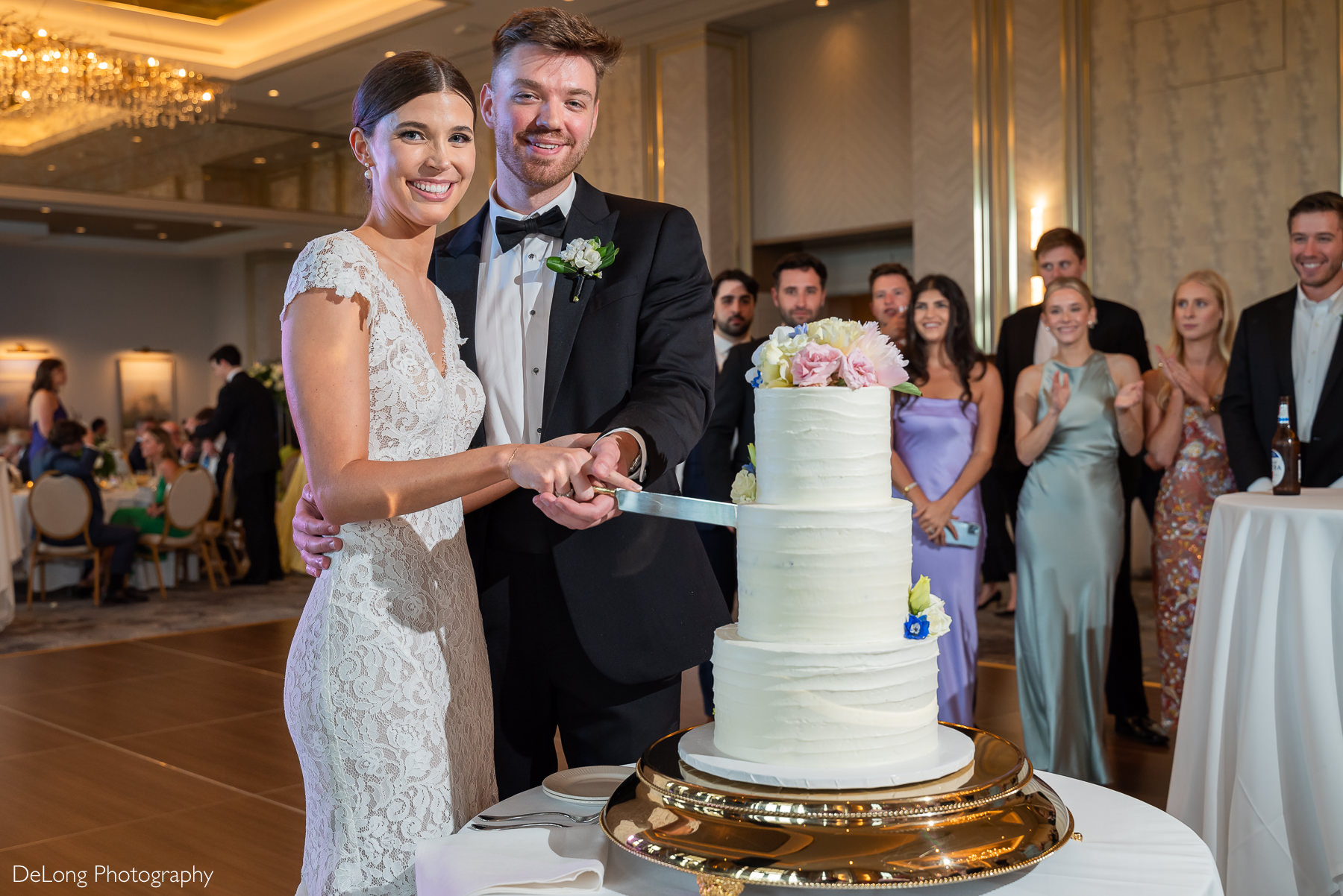 A bride and groom smile at the camera, surrounded by cheerful guests, as they cut their wedding cake inside the Grand Ballroom of Carmel Country Club. Three-tiered white wedding cake is adorned with pink, blue, and white flowers. The bride wears a lace cap-sleeve wedding dress while the grooms looks sharp in a classic black tuxedo. Captured by DeLong Photography.