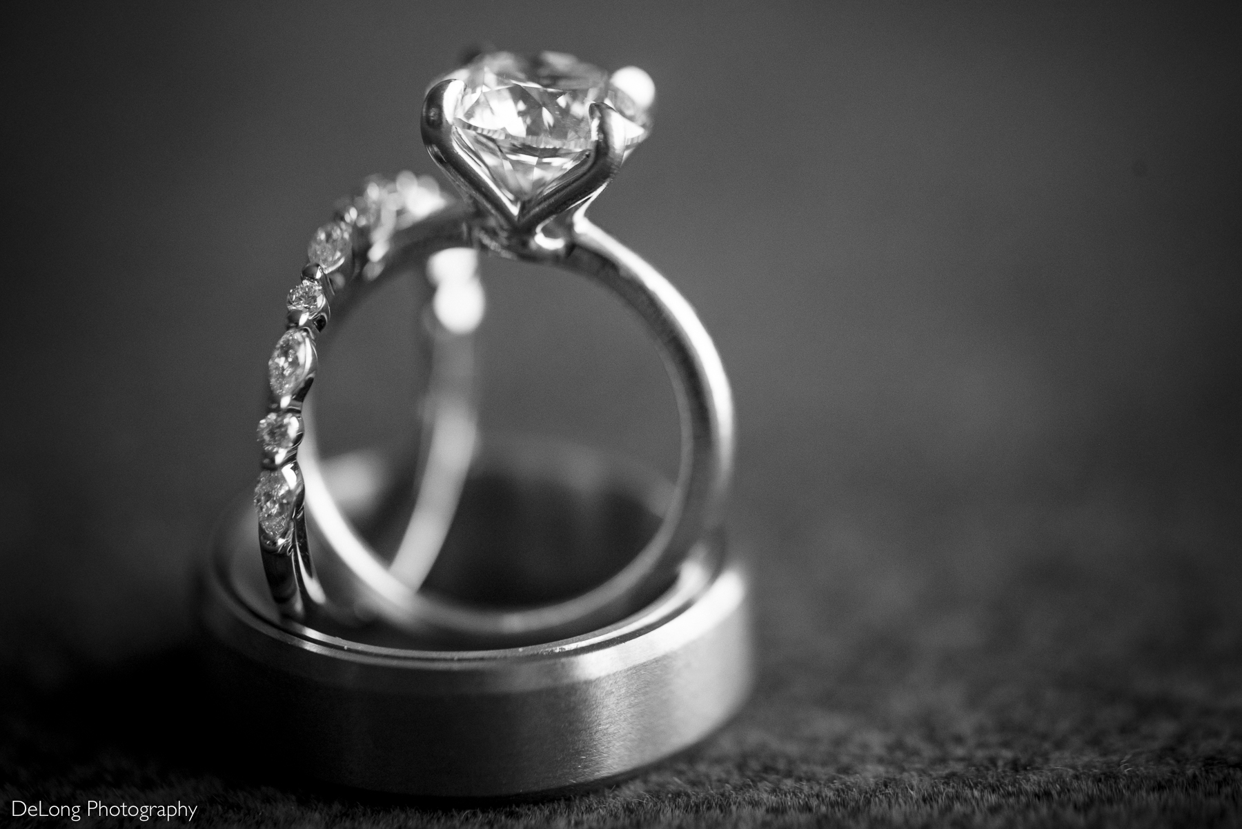 A black and white close-up photograph of three wedding rings stacked together, captured at the Kimpton Tryon Park Hotel. The engagement ring, featuring a sparkling diamond solitaire, sits atop the wedding band with smaller diamonds, while the groom’s simple, smooth band rests beneath them. The image highlights the intricate details of the rings against a softly blurred background. Photo by DeLong Photography.