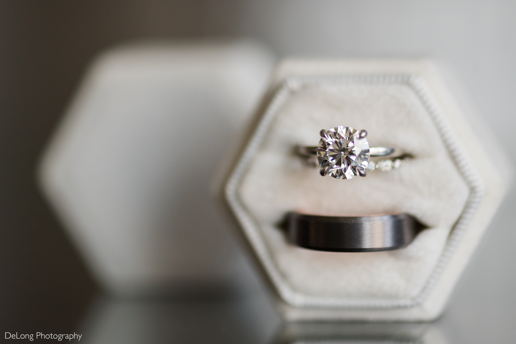 A close-up of a sparkling solitaire engagement ring and a sleek black wedding band, elegantly displayed in a hexagonal velvet ring box. Captured by DeLong Photography at the Kimpton Tryon Park Hotel.