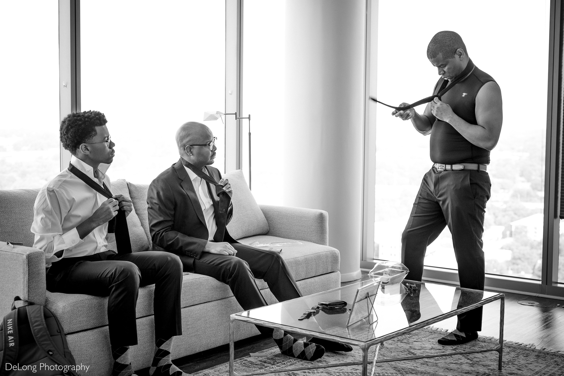 A groom and his two sons, serving as his groomsmen, teaches his sons how to tie their ties in a sleek Uptown Charlotte condo, with a bright cityscape in the background. Captured in black and white, this candid moment highlights their bond and the excitement of the day ahead.
