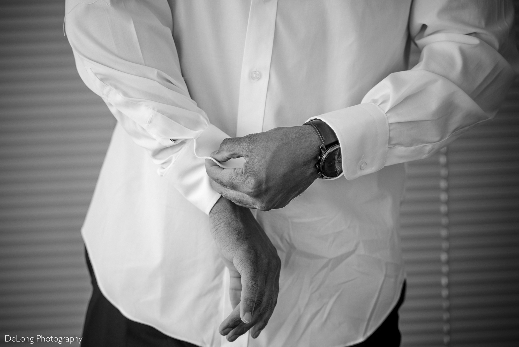 Close-up black-and-white image of a groom fastening the cuff of his crisp white dress shirt. He wears a stylish wristwatch, and his hands are the focal point of the shot. Captured by DeLong Photography at a condo in Uptown Charlotte.