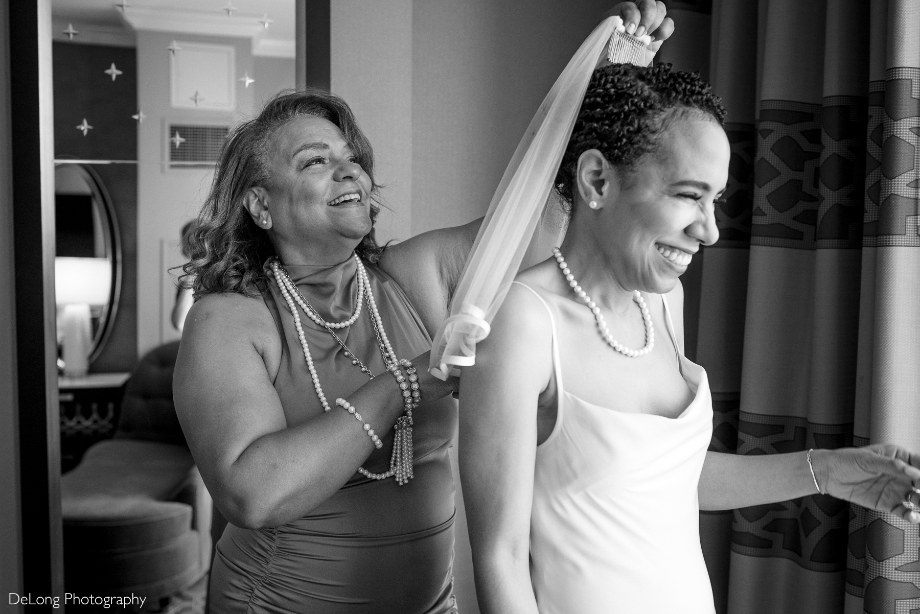 Black and white photograph of a joyful bride laughing as her mother places a veil in her hair. Captured at the Kimpton Tryon Park Hotel by DeLong Photography.