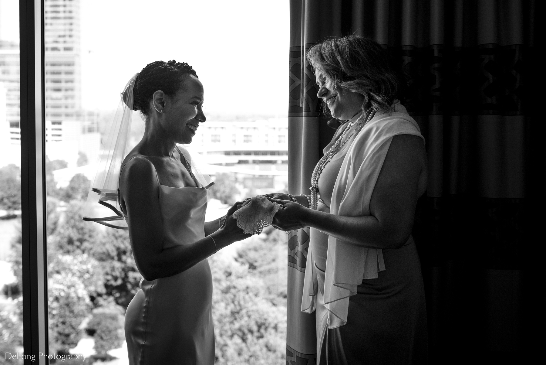 A heartfelt black and white moment between a bride and her mother as they exchange a delicate lace heirloom handkerchief. They smile warmly at each other near a window overlooking the cityscape. Captured at the Kimpton Tryon Park Hotel by DeLong Photography.
