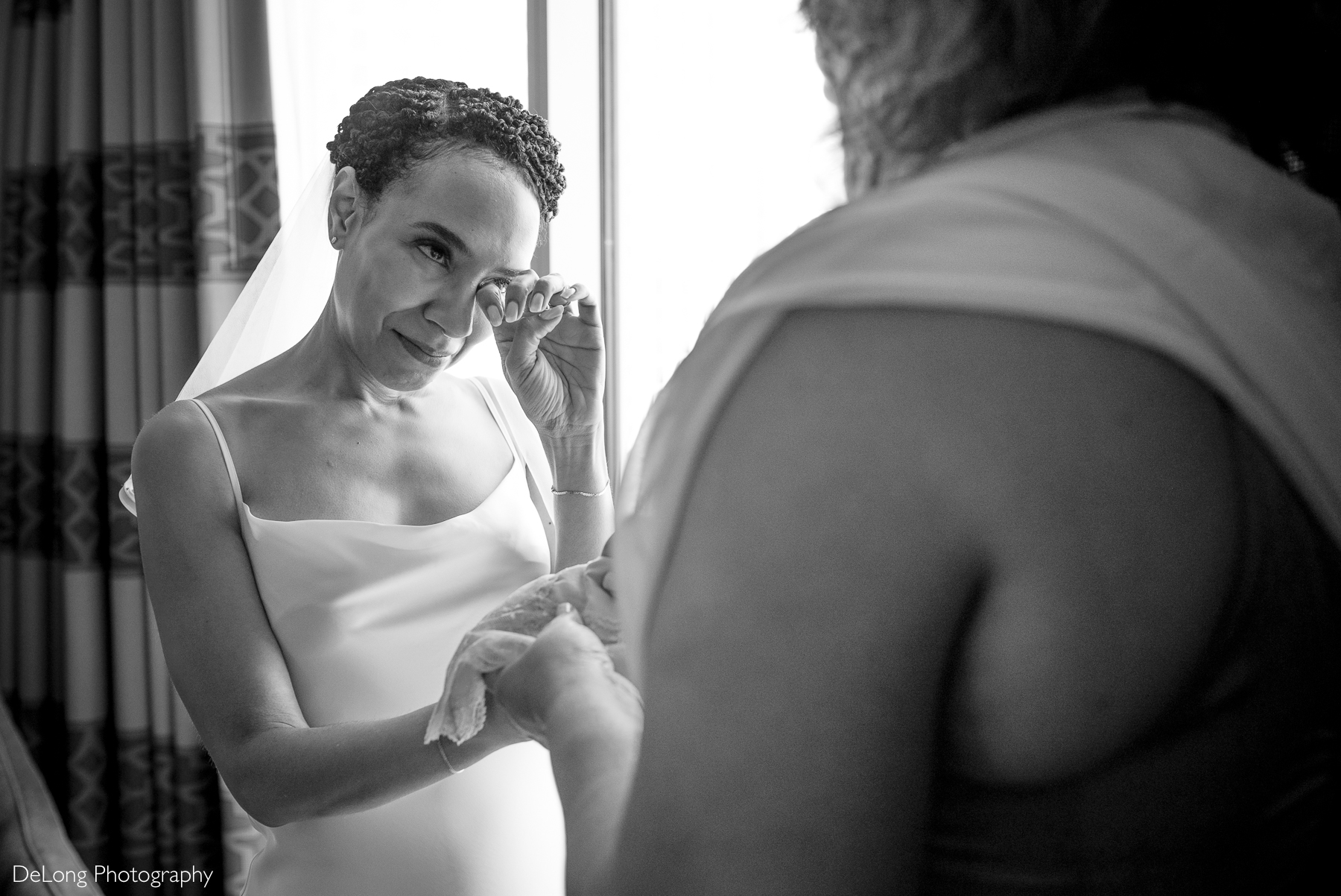 A bride wipes away a tear as she shares an emotional moment with her mother, who offers her a delicate lace handkerchief that is a family heirloom. The black and white image captures their deep bond in a softly lit room at the Kimpton Tryon Park Hotel. Captured by DeLong Photography.