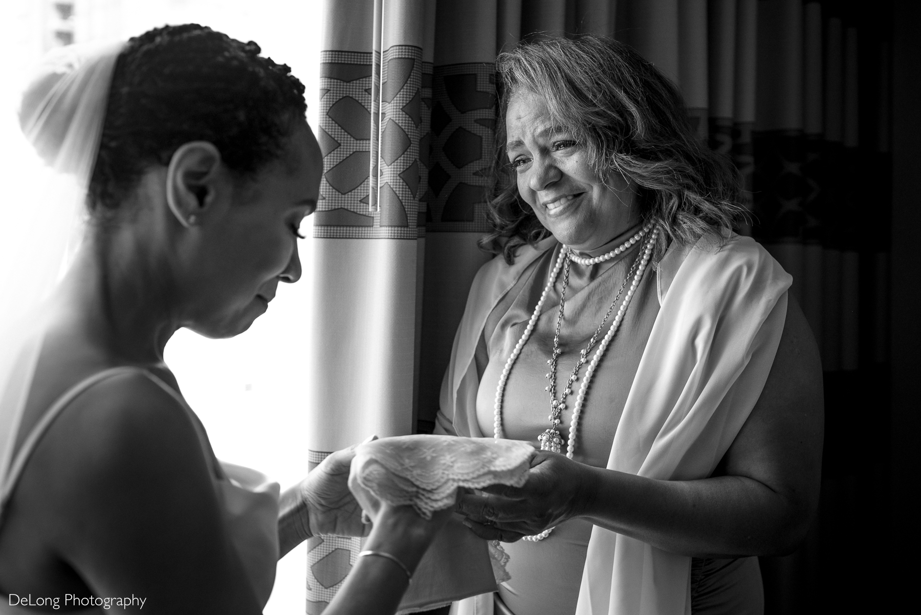 A heartfelt black and white moment between a bride and her mother at Kimpton Tryon Park Hotel in Charlotte, NC. The bride, wearing a delicate veil, looks down with emotion as she receives a lace handkerchief that is a family heirloom. The woman, dressed elegantly with layered pearl necklaces, smiles warmly, conveying love and support. Soft natural light filters through the window, enhancing the intimate and sentimental atmosphere. Captured by DeLong Photography.