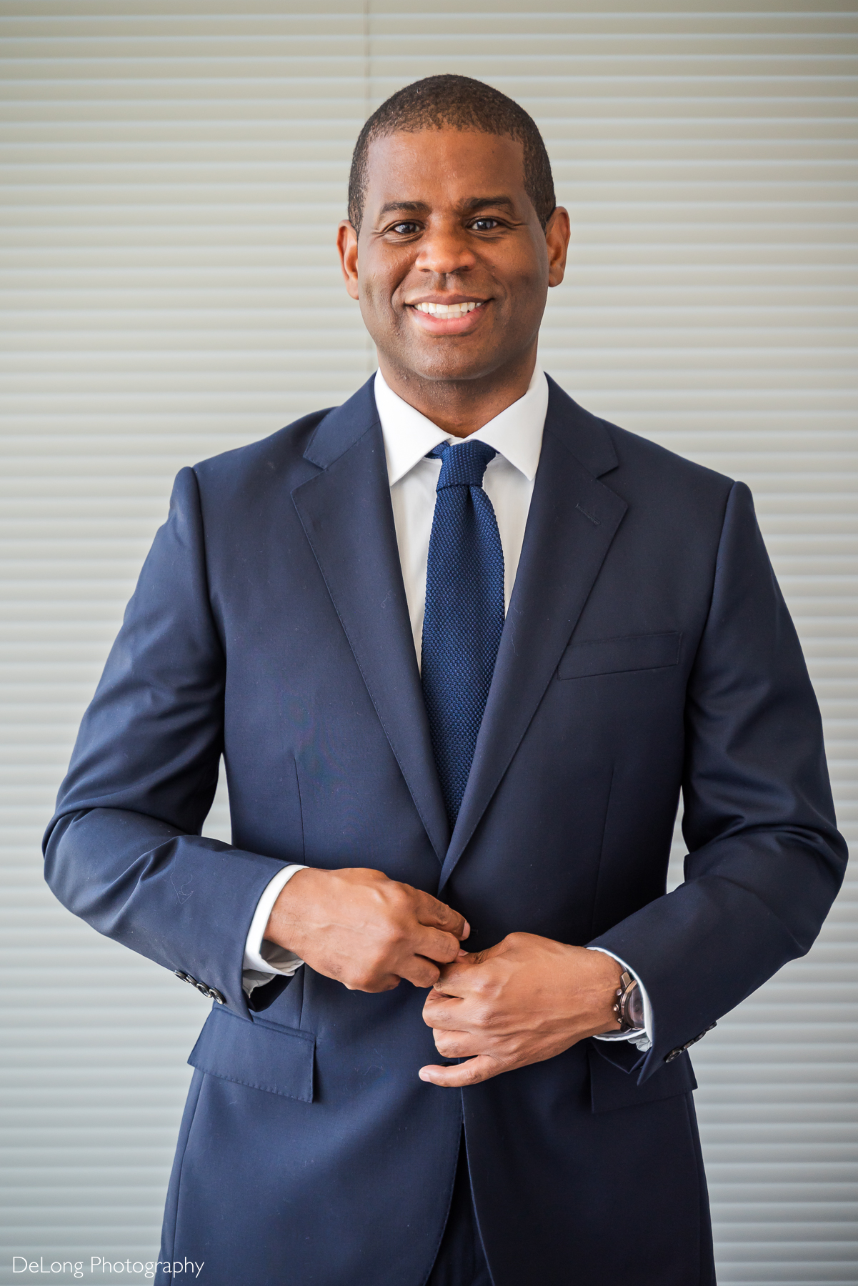 A confident groom in a navy blue suit and tie buttons his jacket while smiling warmly at the camera. Captured at his condo in Uptown Charlotte, the modern, neutral-toned background emphasizes his polished and sophisticated look. The natural lighting highlights his features, creating a professional yet relaxed portrait. Taken by DeLong Photography.