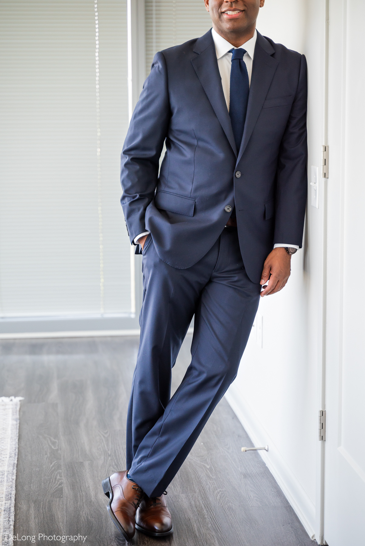 A groom dressed in a navy blue suit, white dress shirt, and dark blue tie leans casually against a wall in his modern Uptown Charlotte condo. He wears polished brown dress shoes and a wristwatch, exuding confidence and elegance. Captured by DeLong Photography.