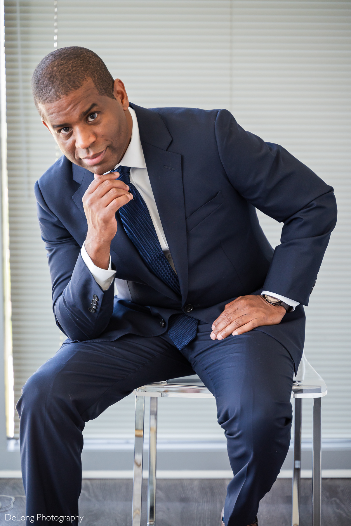 A groom in a navy blue suit, white dress shirt, and dark blue tie sits confidently on a modern clear acrylic stool in his Uptown Charlotte condo. He leans slightly forward, resting one hand on his knee and the other near his chin, giving a relaxed yet poised expression. Captured by DeLong Photography.
