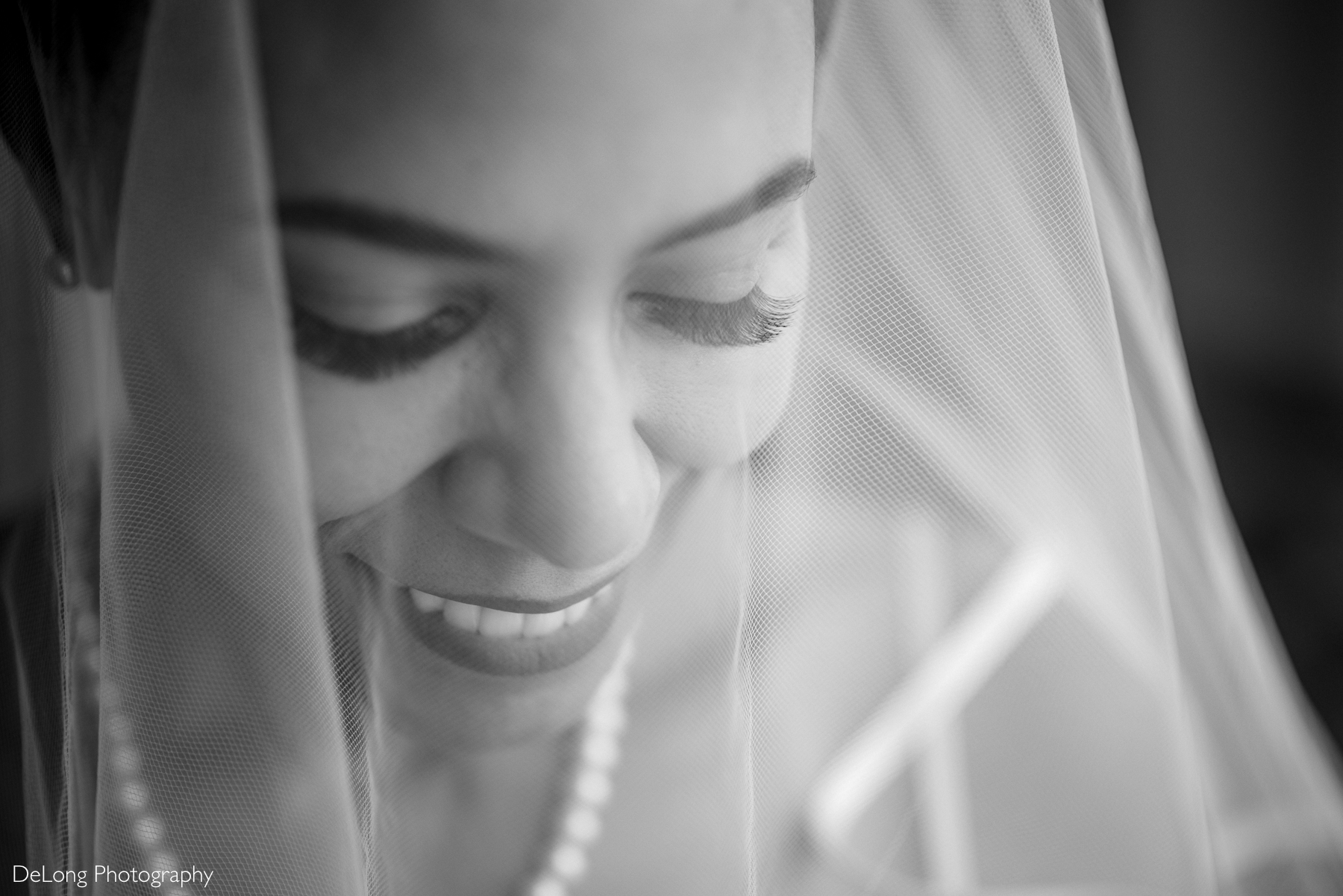 A black and white close-up portrait of a bride smiling softly beneath her veil. Her eyes are lowered, and her long lashes frame her serene expression. She wears a pearl necklace, and soft natural light highlights her features. Captured at Kimpton Tryon Park Hotel in Charlotte, NC, by DeLong Photography.