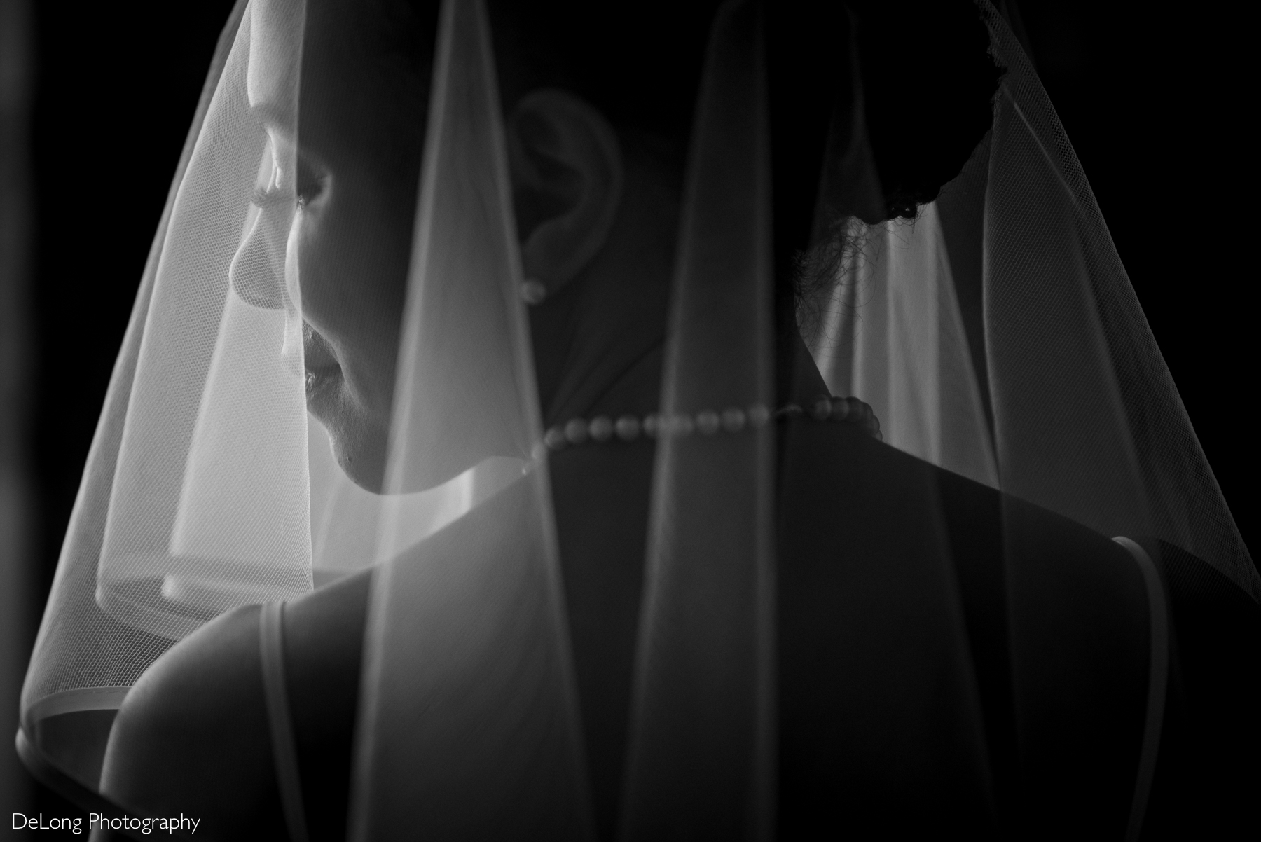 A black and white portrait of a bride captured from behind, with her face softly illuminated as she smiles beneath her veil. The delicate fabric drapes over her shoulders, highlighting the elegance of her pearl necklace. Taken at Kimpton Tryon Park Hotel in Charlotte, NC, by DeLong Photography.