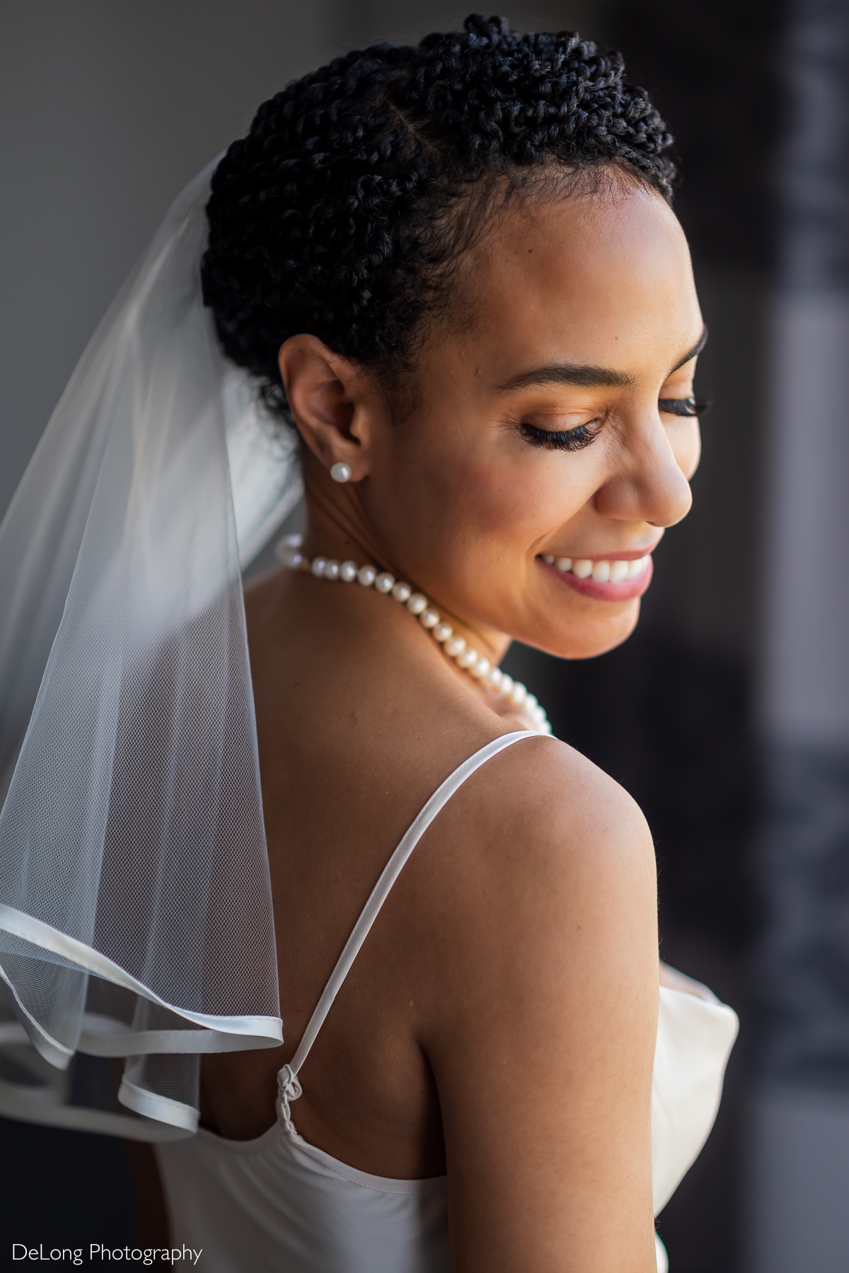 A radiant bride with short, textured hair smiles softly as she looks over her shoulder, her pearl necklace and delicate veil adding timeless elegance. The natural light highlights her glowing complexion and joyful expression. Captured at Kimpton Tryon Park Hotel in Charlotte, NC, by DeLong Photography.