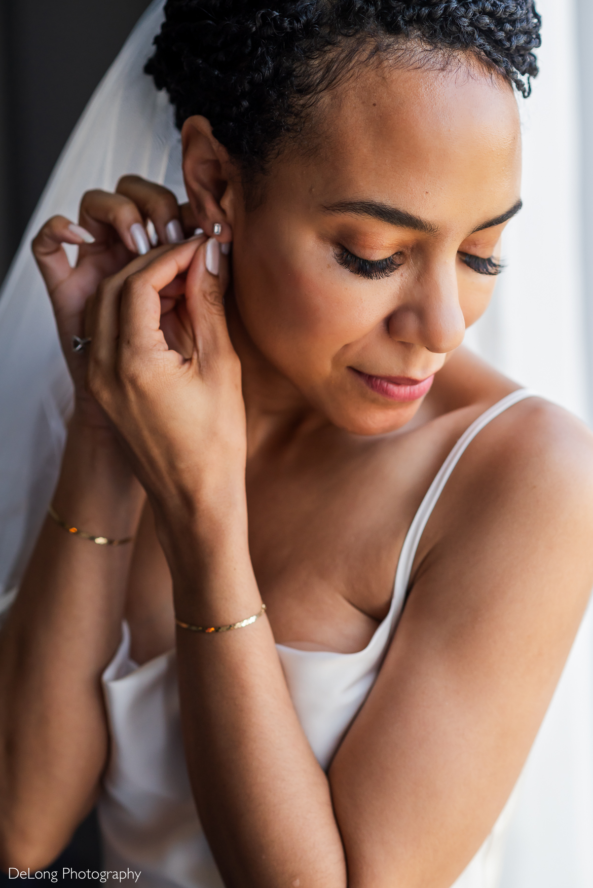 A bride with short, textured hair and a sheer white veil gently adjusts her stud earring while looking down with a soft smile. She wears a minimalist white gown, gold bracelets, and a delicate engagement ring. Her makeup is natural, highlighting her long eyelashes and radiant complexion. Soft natural light from a nearby window illuminates her face. The photo was taken at the Kimpton Tryon Park Hotel. Captured by DeLong Photography.