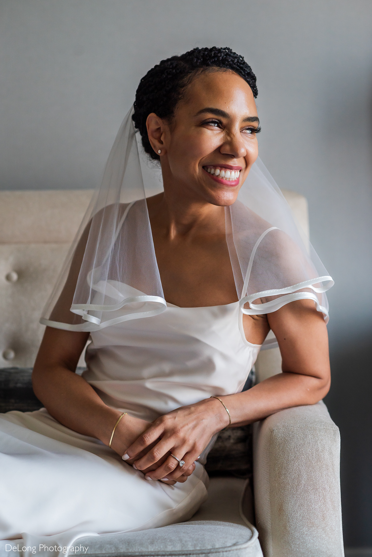 A radiant bride with a joyful smile sits on a plush, neutral-toned armchair at the Kimpton Tryon Park Hotel. She wears a modern white gown with delicate straps and a short, sheer veil with satin trim. Her hands, adorned with a sparkling engagement ring and gold bracelets, rest gently on her lap. Soft natural light highlights her glowing complexion as she looks off to the side with happiness. Captured by DeLong Photography.