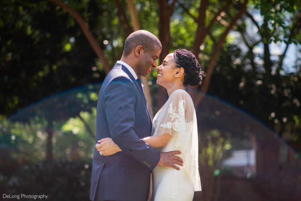 Bride and groom embrace in Romare Bearden Park, smiling with noses touching. Sun flare creates a soft arch at the bottom of the image.