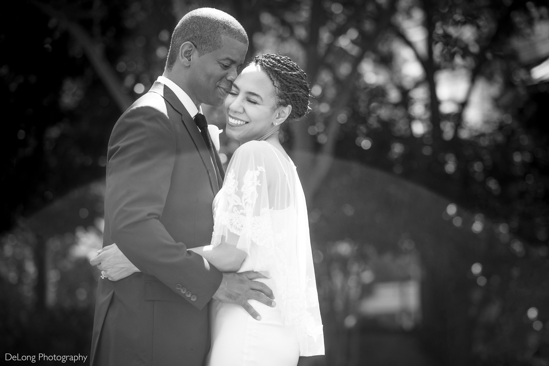 A joyful Black couple embraces on their wedding day at Romare Bearden Park. The groom, wearing a classic navy suit, gently holds his bride, who smiles radiantly in a modern white gown with delicate lace details. Sunlight filters through the trees, creating a dreamy, romantic atmosphere. Captured by DeLong Photography.