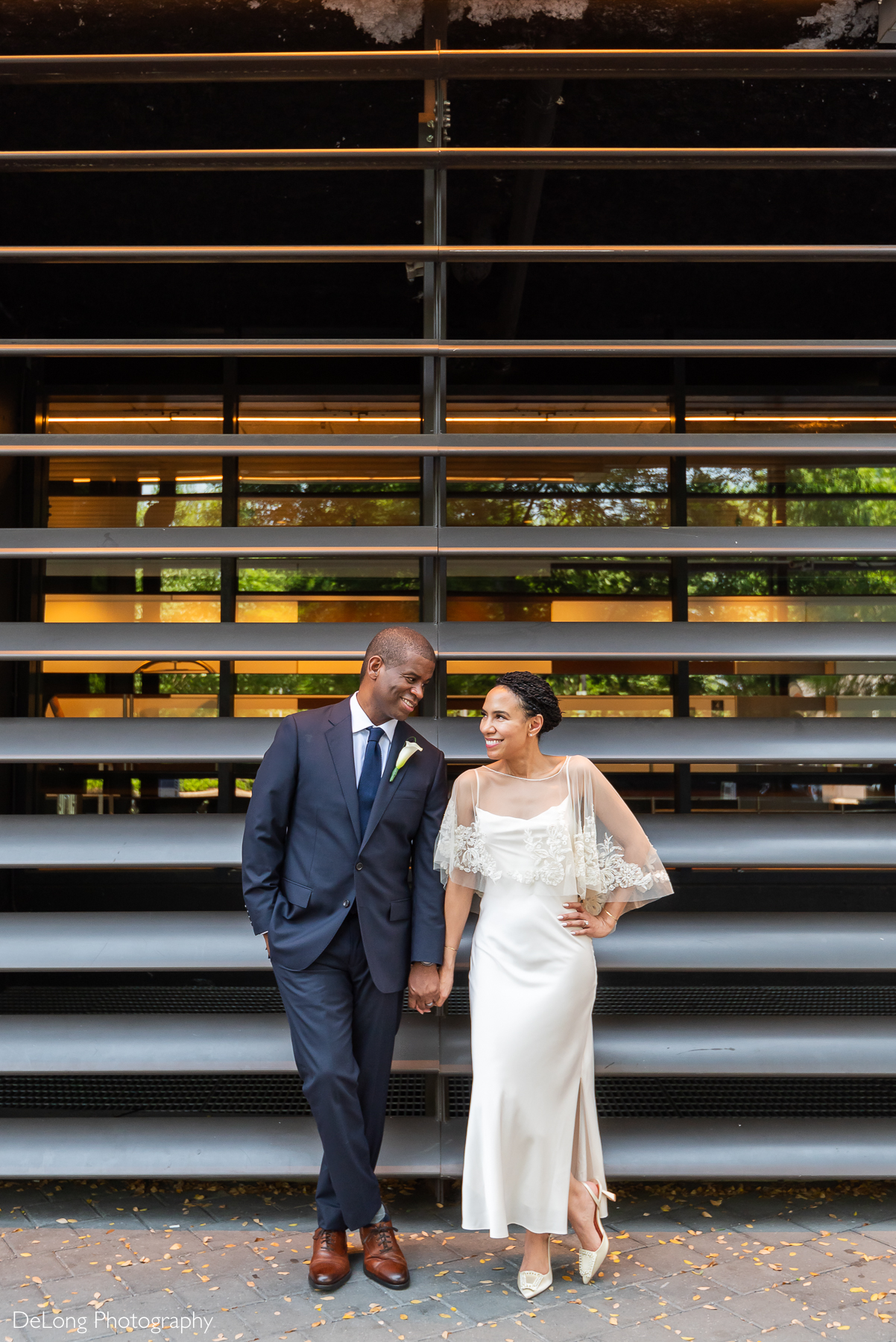 A stylish newlywed couple shares a joyful moment outside the Kimpton Tryon Park Hotel near Romare Bearden Park. The groom, in a navy suit, and the bride, in a chic white gown with lace sleeves, hold hands and smile at each other against a modern architectural backdrop. Captured by DeLong Photography.