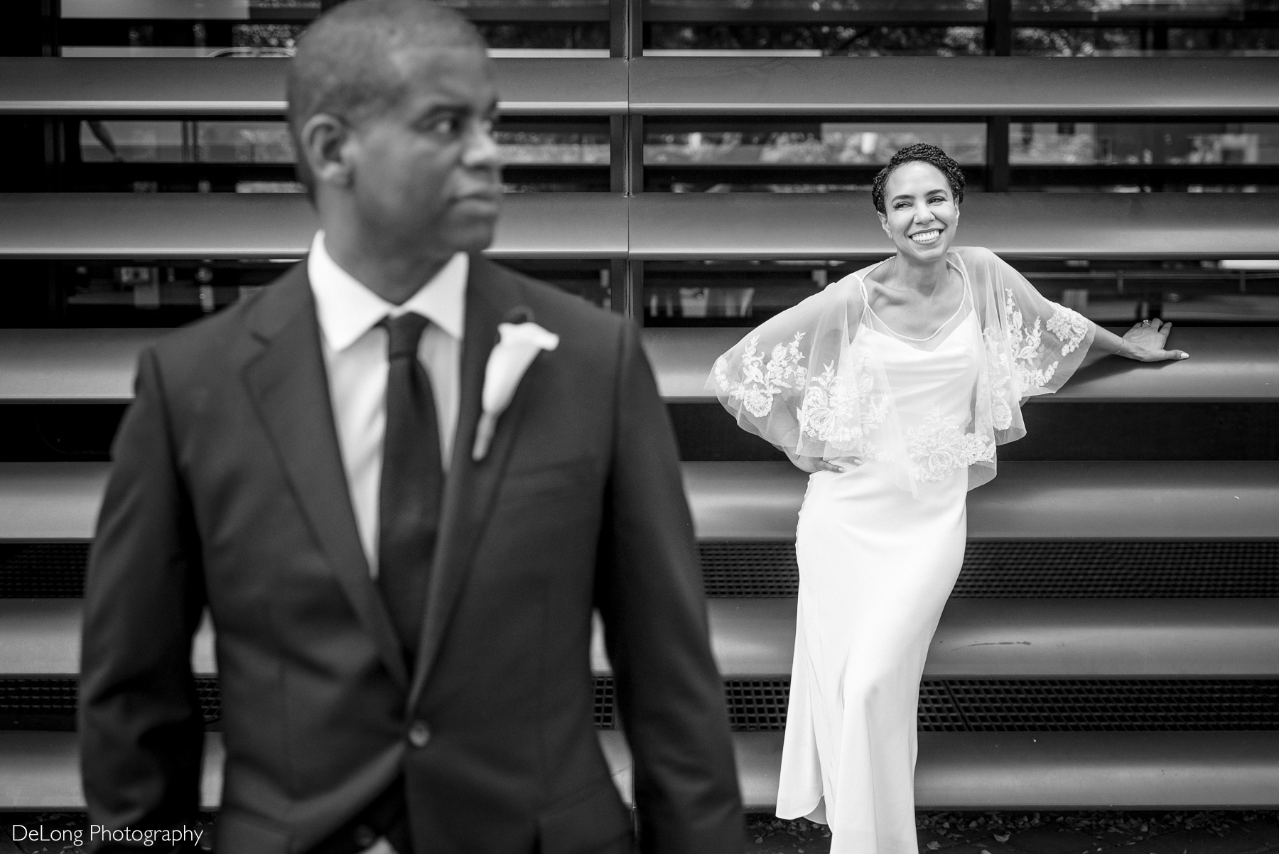 A black and white photograph of a newlywed couple taken outside the Kimpton Tryon Park Hotel near Romare Bearden Park. The groom, dressed in a sharp black suit with a white boutonniere, stands in the foreground looking off to the side. In the background, the bride leans playfully against a modern metal structure, smiling radiantly in her elegant satin gown with delicate lace cape sleeves. Captured by DeLong Photography.